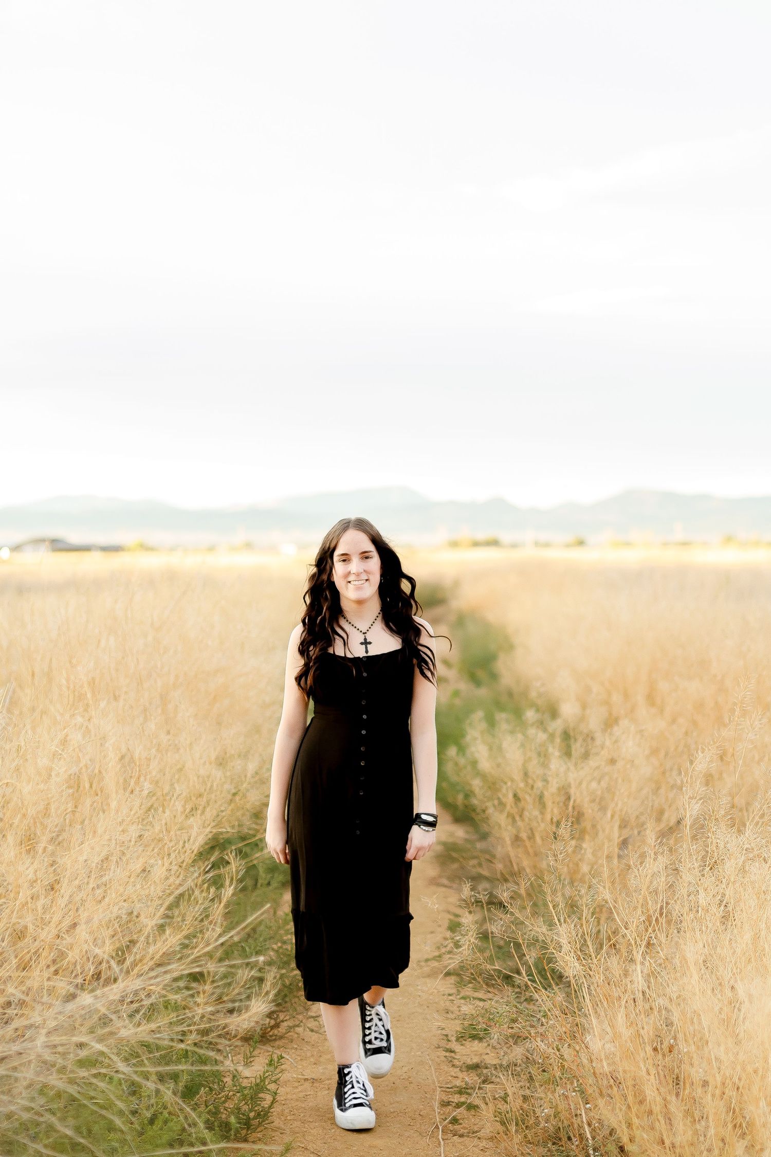 Young woman in black midi dress and sneakers walking along a dirt path through golden wheat fields.