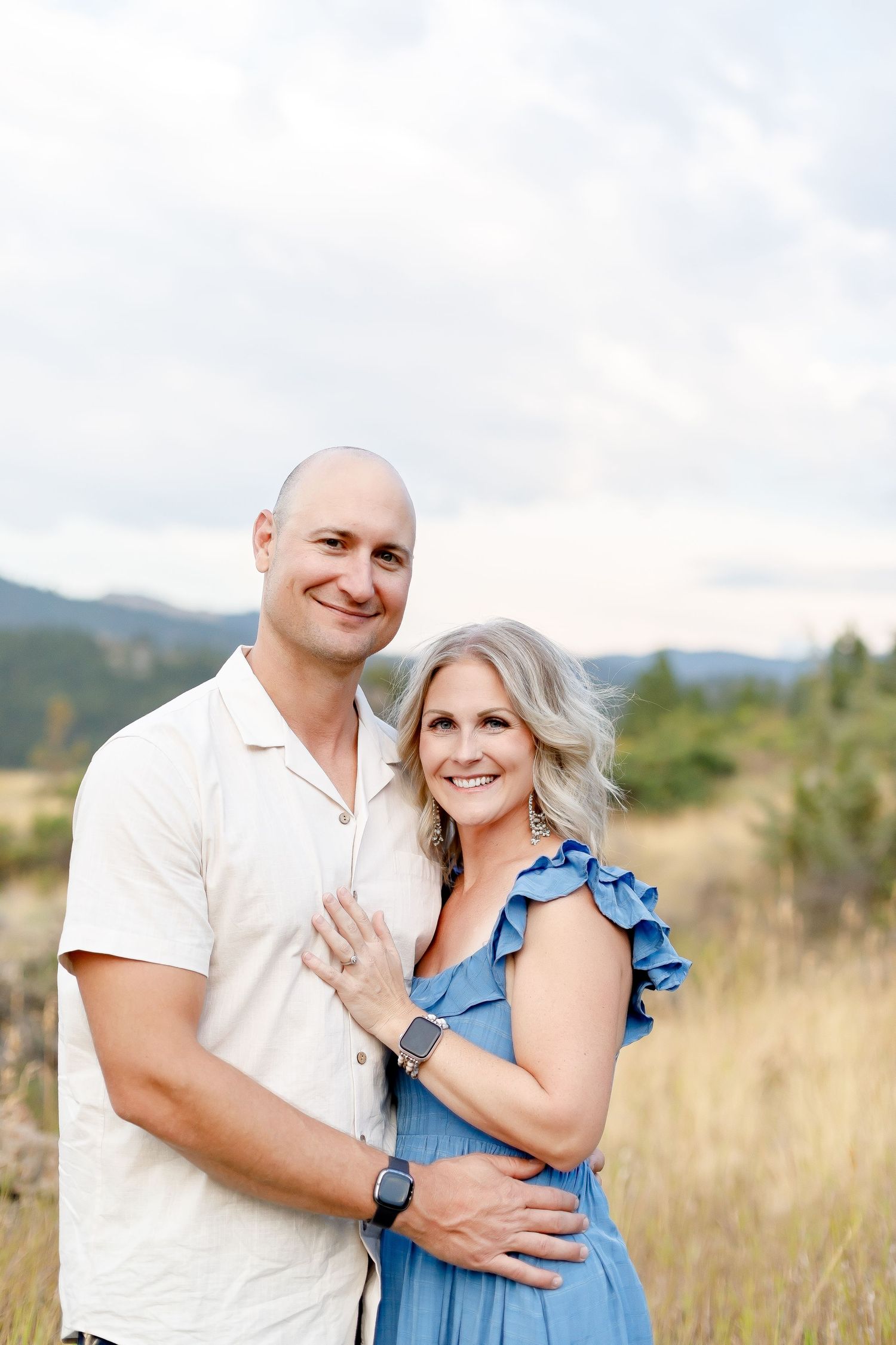 A couple smiling together in a mountain meadow, woman in a blue ruffled dress and man in white.
