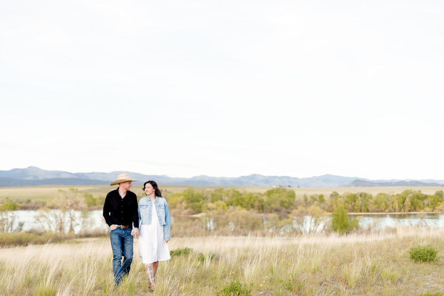 Couple walking hand in hand through a grassy field with a lake and mountains behind.