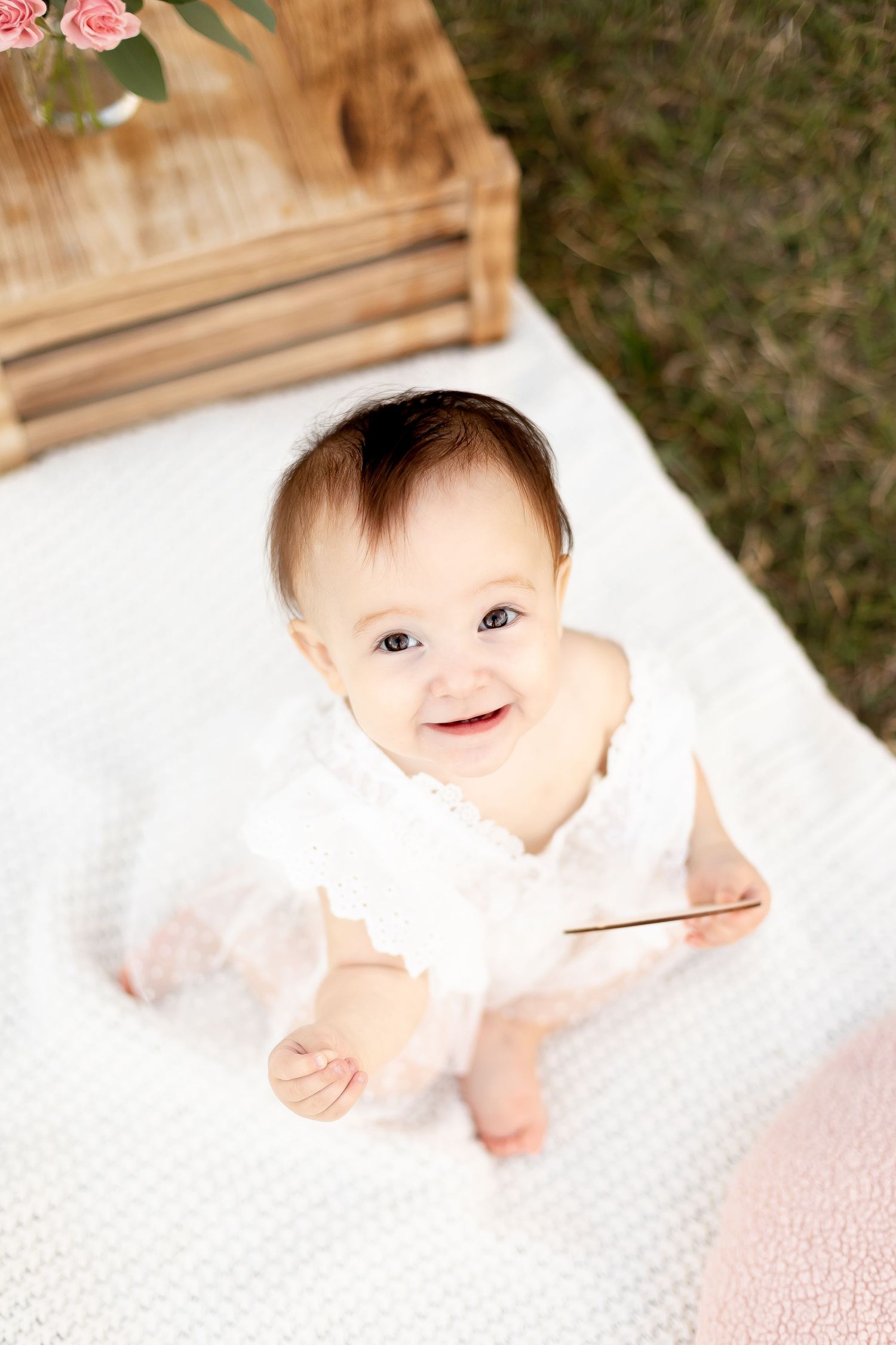 Smiling baby girl in white lace dress sitting on blanket outdoors at one year birthday photo session.