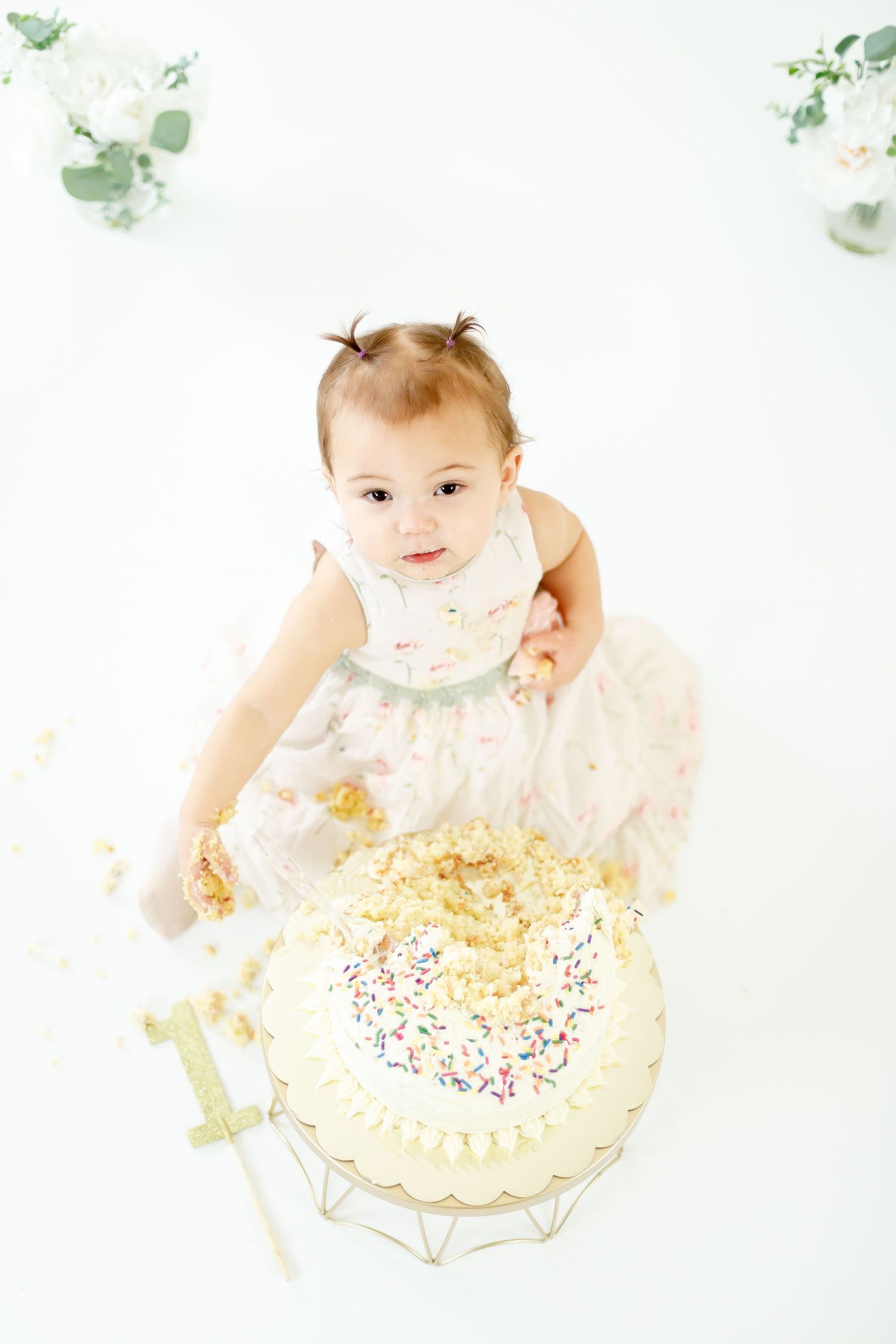 Overhead view of a baby girl in a floral dress smashing her first birthday cake with sprinkles.