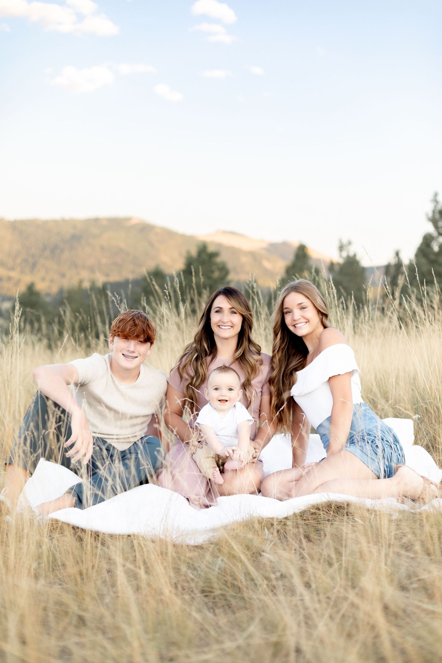 A mother sits in a golden field with her two teens and baby on a white blanket outdoors.