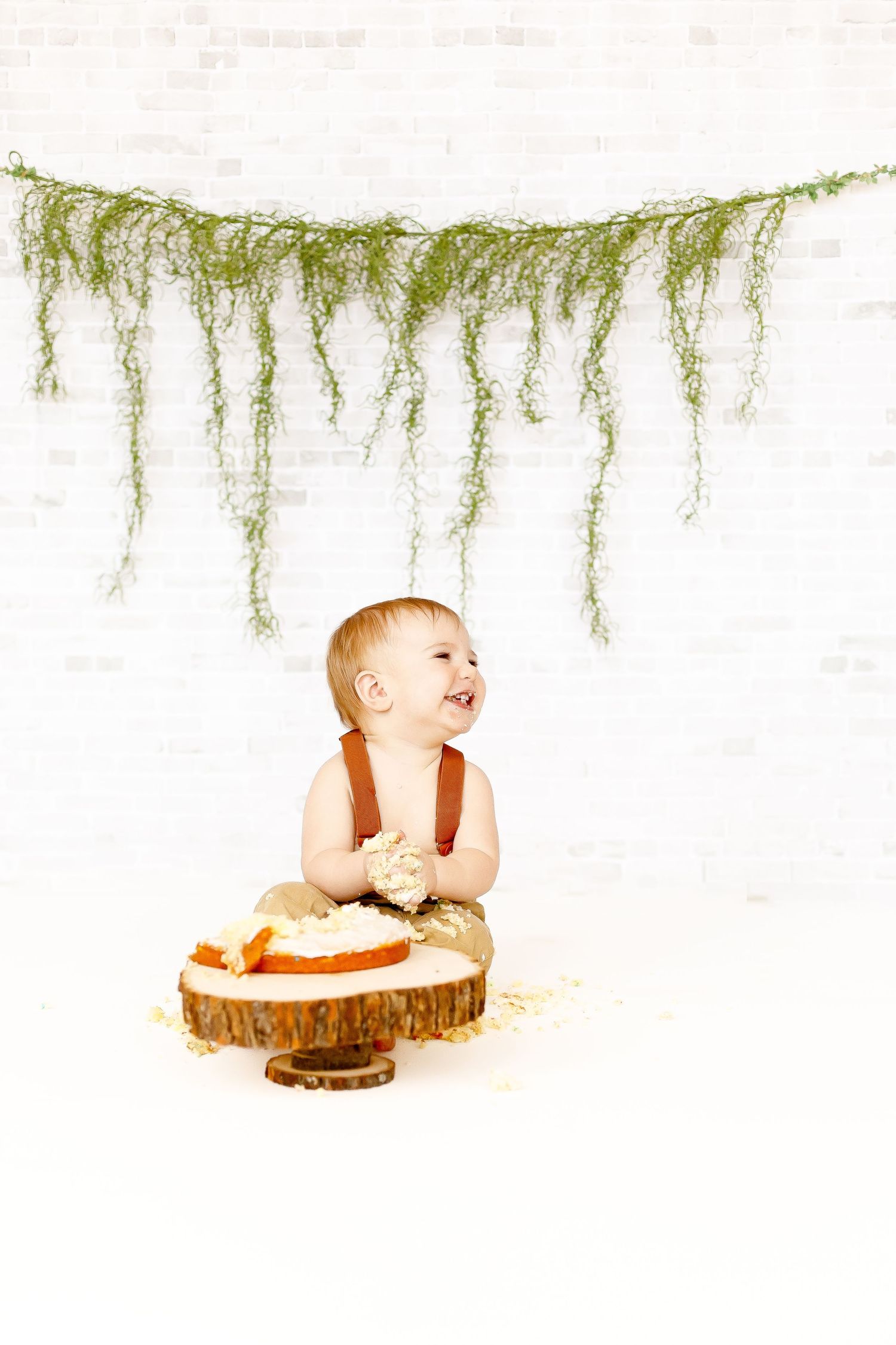 A laughing baby boy in tan overalls and suspenders sits next to a smashed birthday cake indoors.