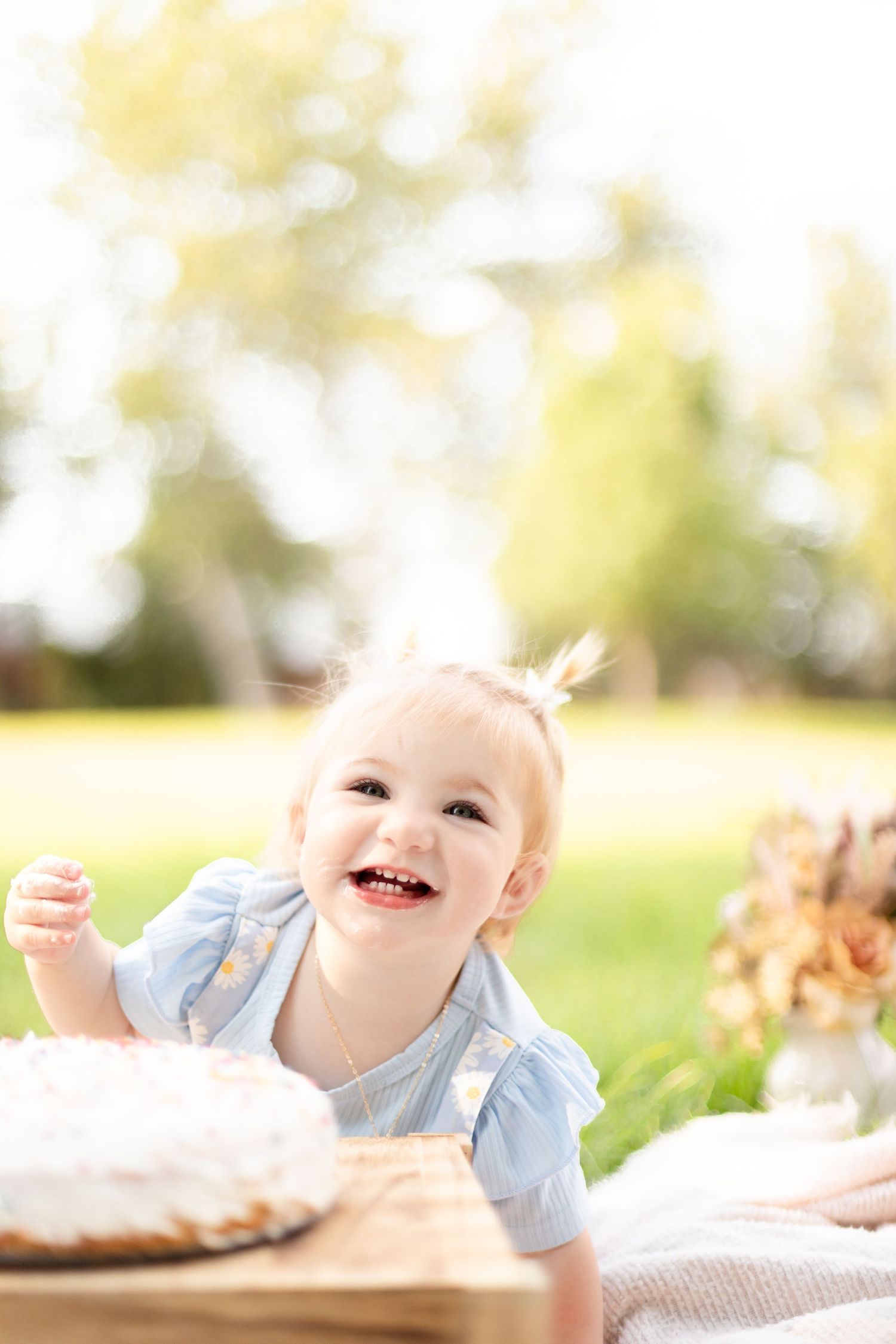 Happy baby girl giggling near her white smash cake on a wooden crate during an outdoor first birthday session.