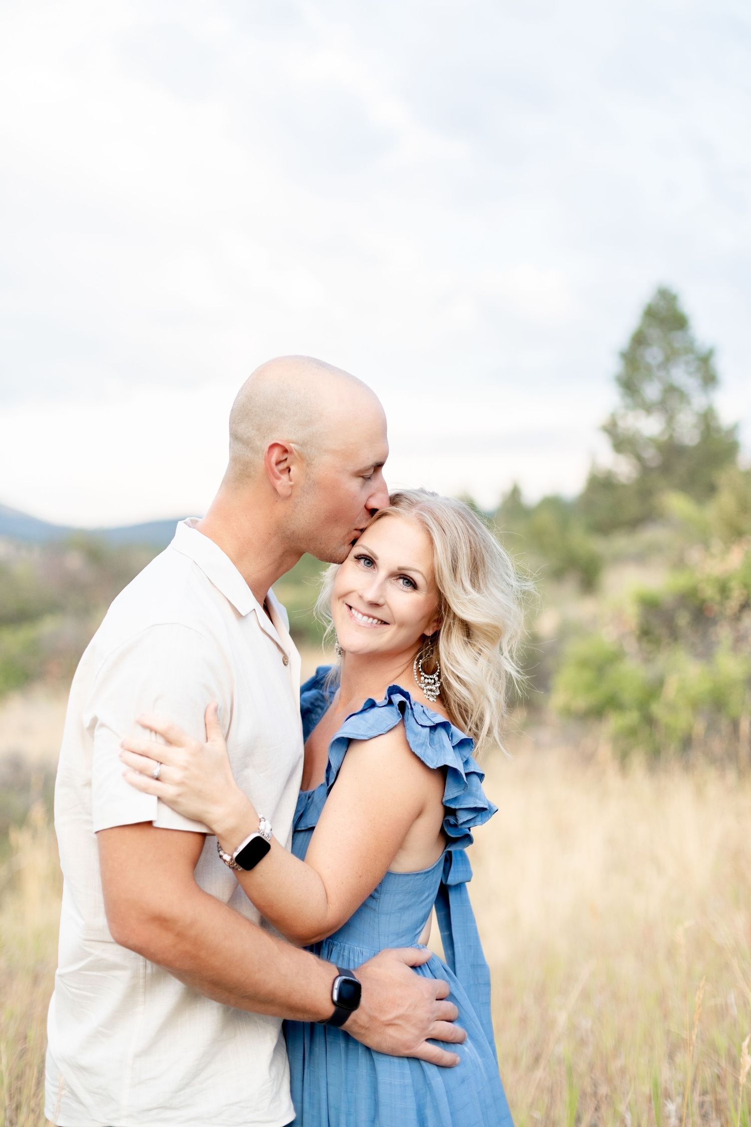 Couple embracing in a field, man kissing woman's forehead, woman in blue dress smiling.