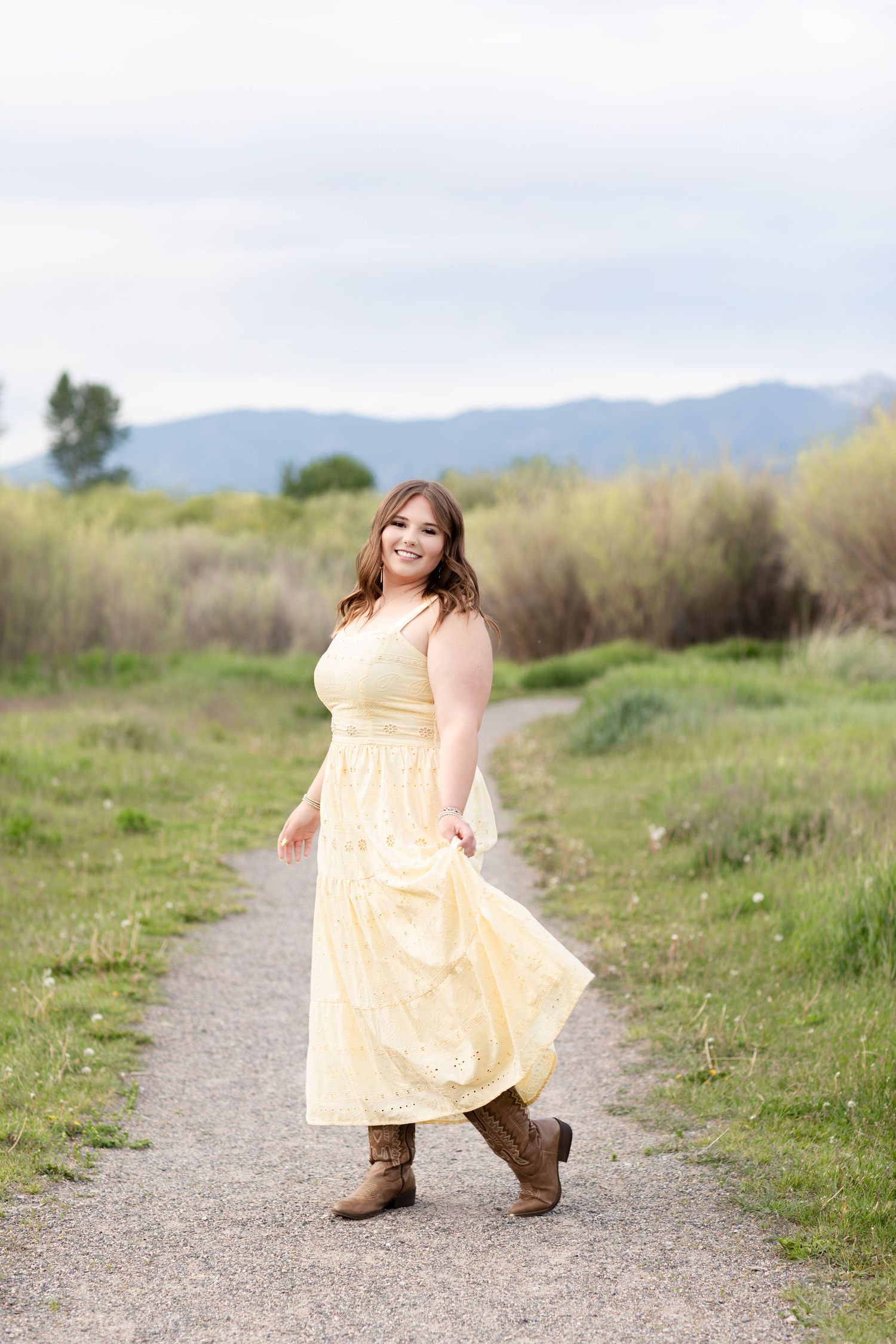 Smiling young woman in yellow maxi dress and cowboy boots twirling on a gravel path with mountains.
