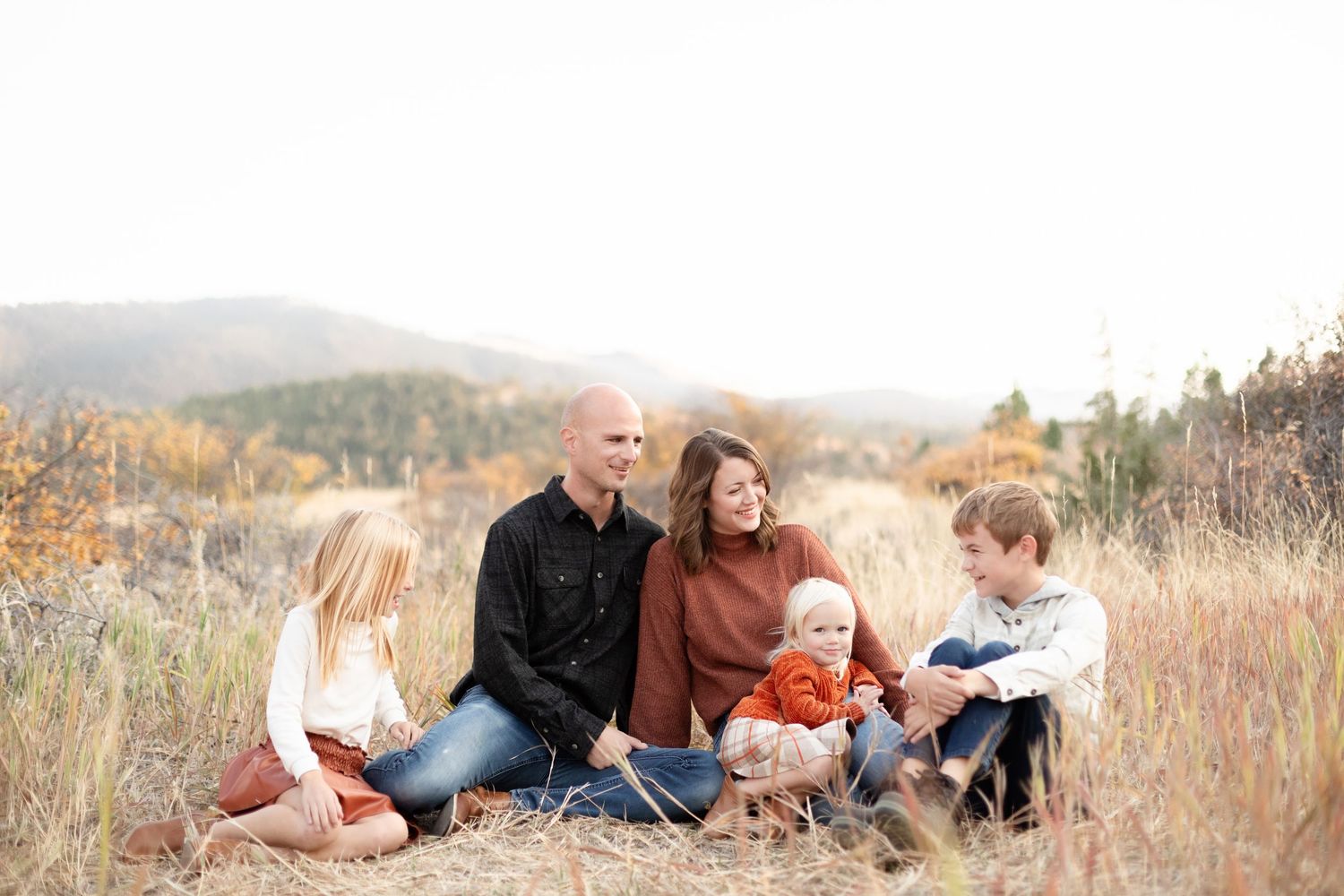 Family of five sitting together in a golden autumn field with mountain backdrop during fall photo session.