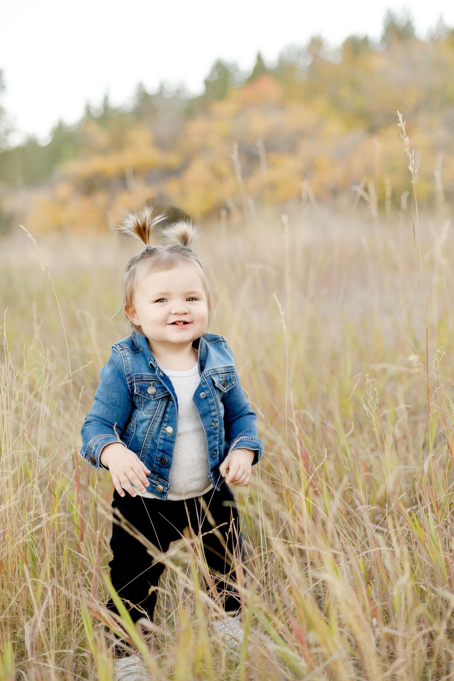 A happy toddler girl in a denim jacket stands smiling in tall golden grass in an autumn field.