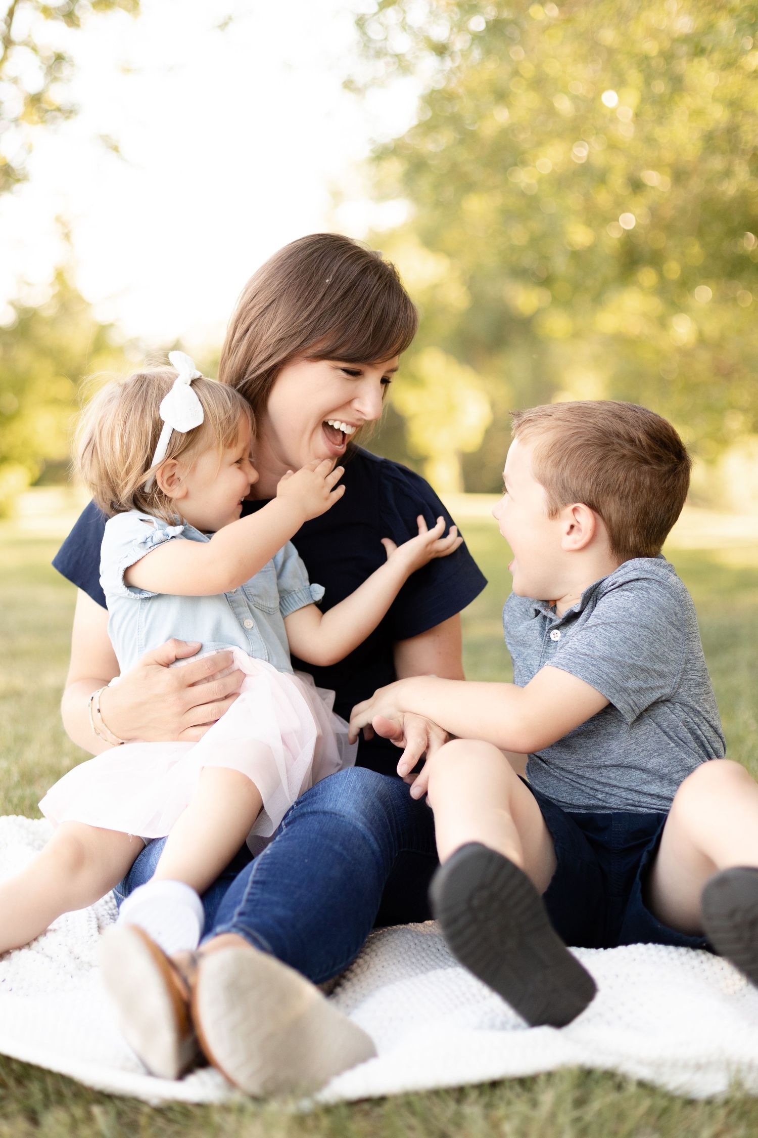 Laughing mother sits on a blanket in the park with her two young children.