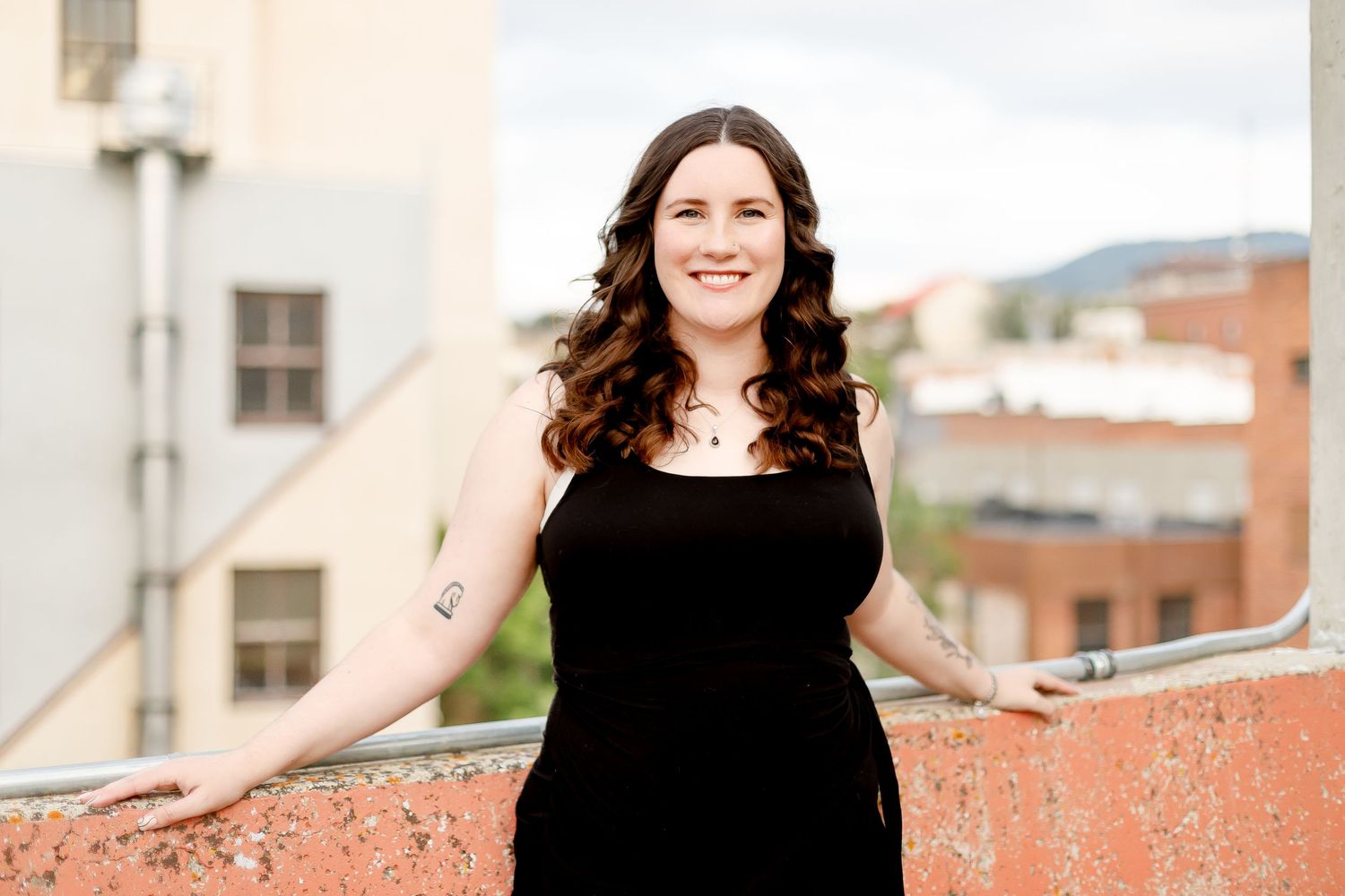 Smiling woman in black dress posing against a red wall with a city skyline in the background.
