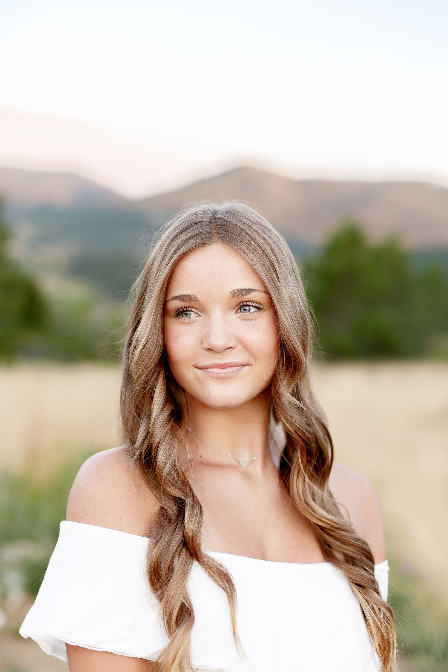 Close-up portrait of young woman in white off-shoulder top with mountain scenery behind her.