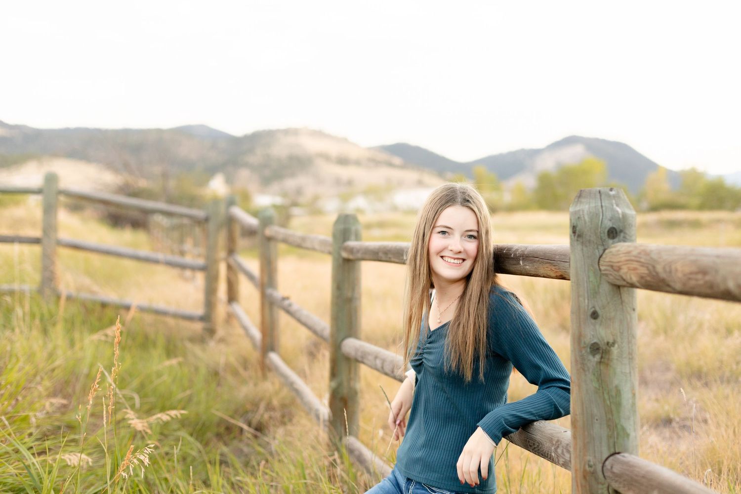 Cheerful teenage girl in teal top leaning on a wooden fence with mountain meadow in the background.