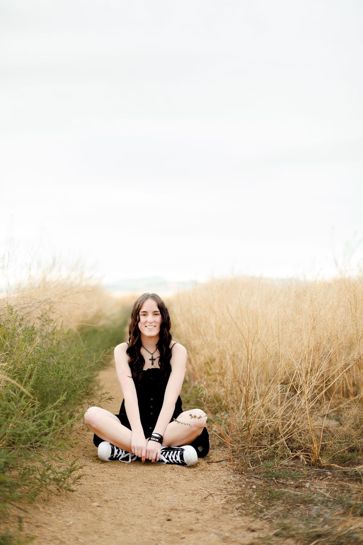 Smiling young woman in black dress sitting cross-legged on a dirt path surrounded by tall golden grass.