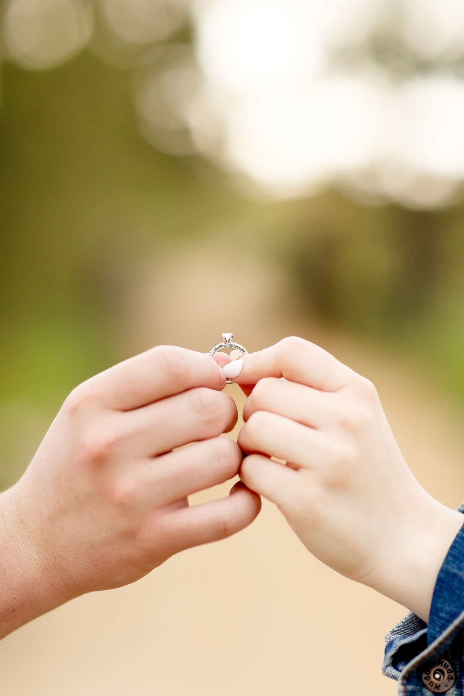 Two hands holding a delicate diamond engagement ring outdoors with a soft bokeh background.