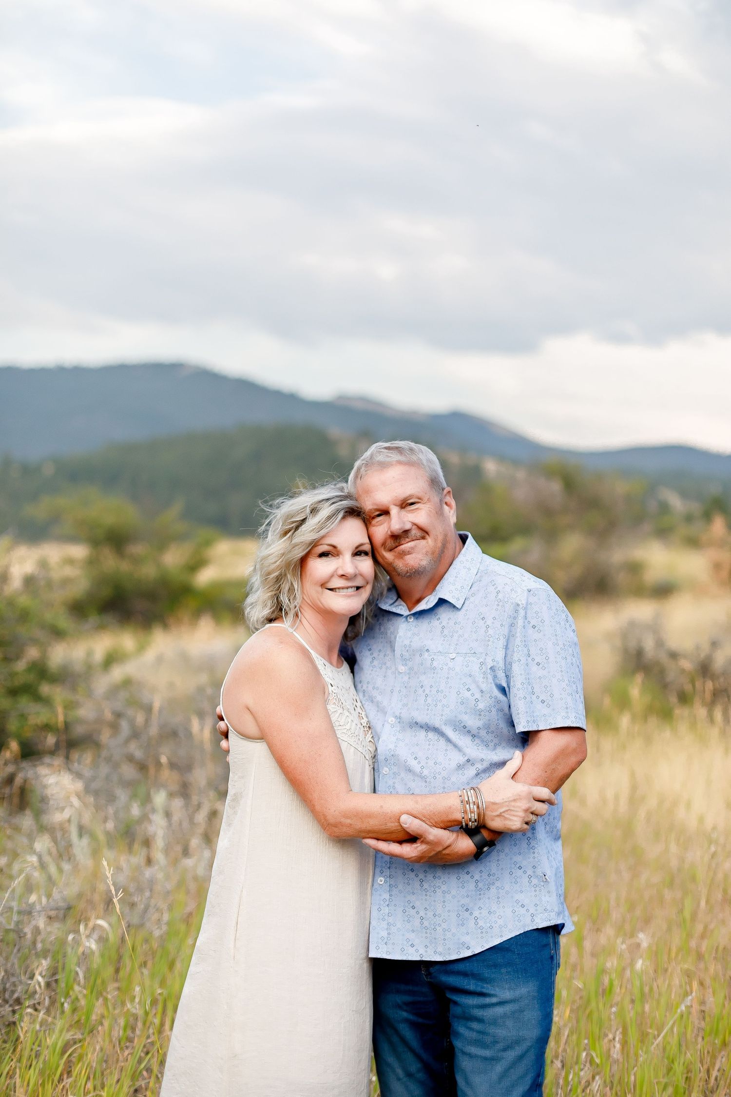 A smiling mature couple embracing in a golden field with mountain scenery in the background.