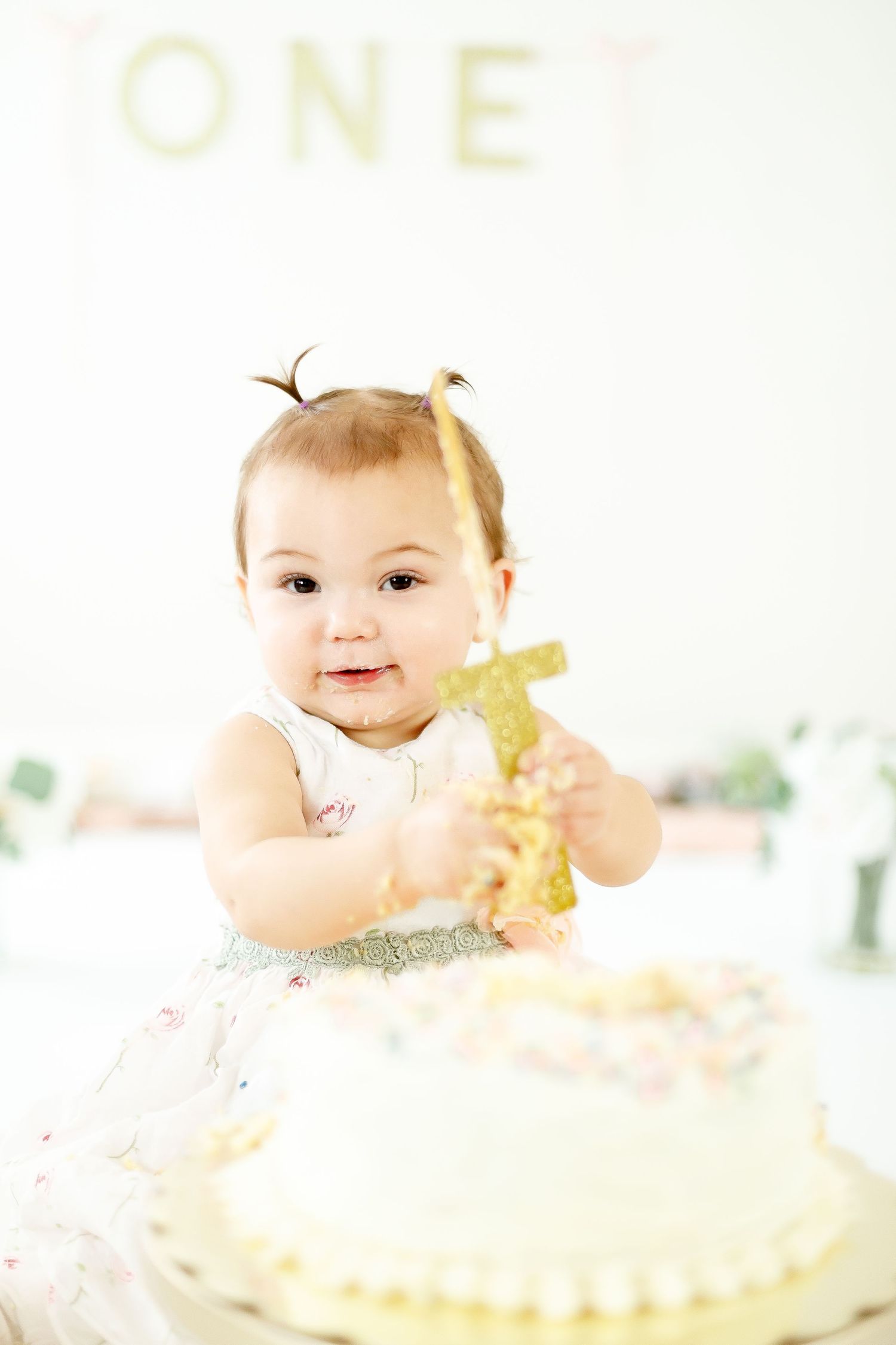 A smiling baby girl holds a gold number one candle during her first birthday cake smash photoshoot.
