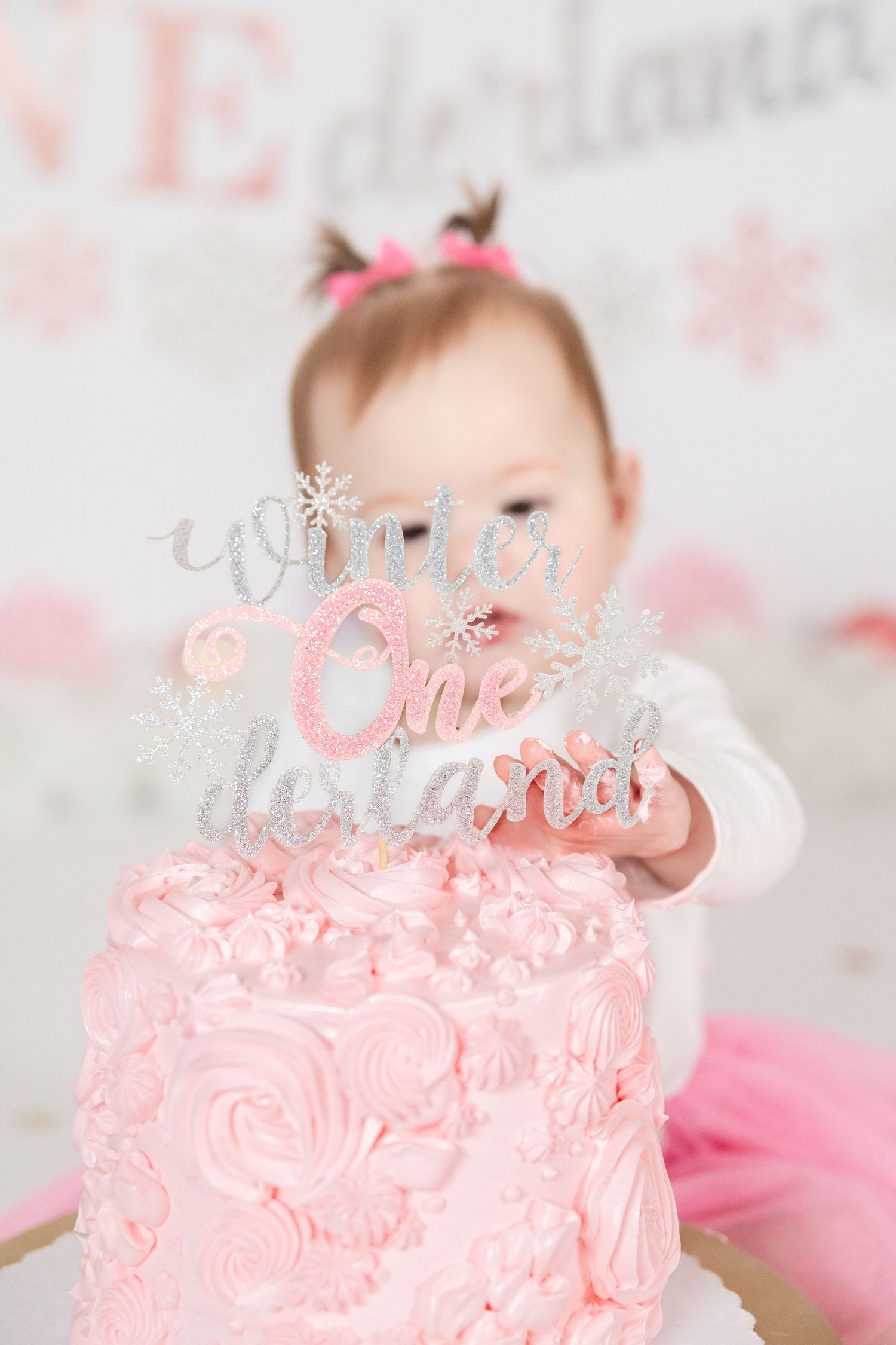 Baby girl reaching for pink Winter ONEderland first birthday smash cake with glitter topper.
