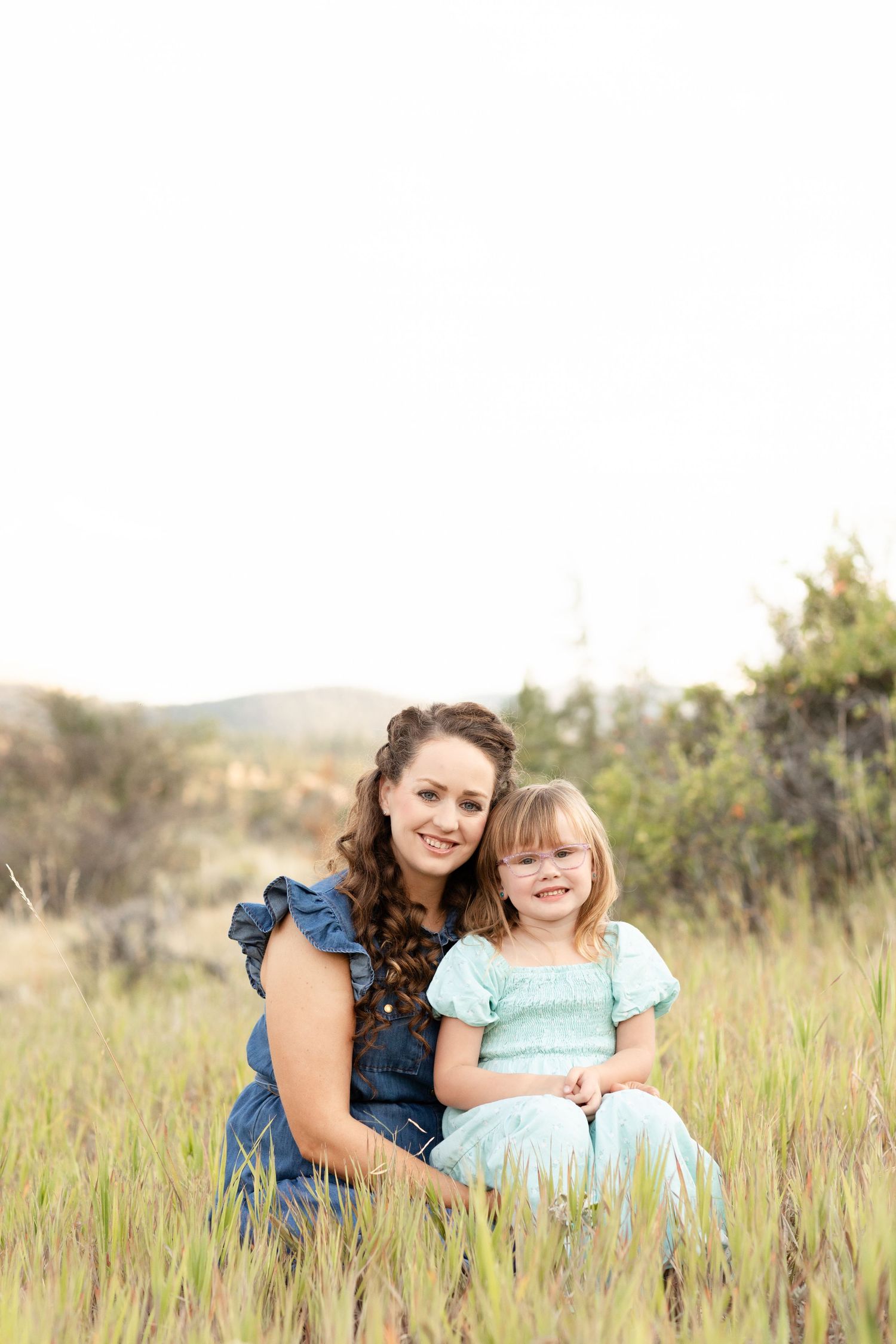 A mother and young daughter smile together in a grassy field during an outdoor family photo session.