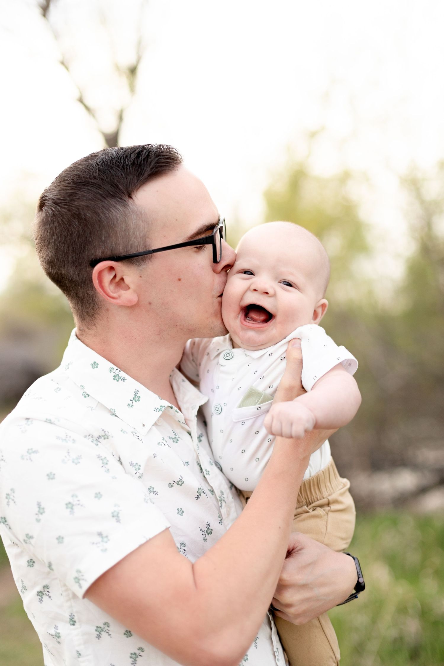Father in floral shirt kissing cheek of giggling baby outdoors in nature.