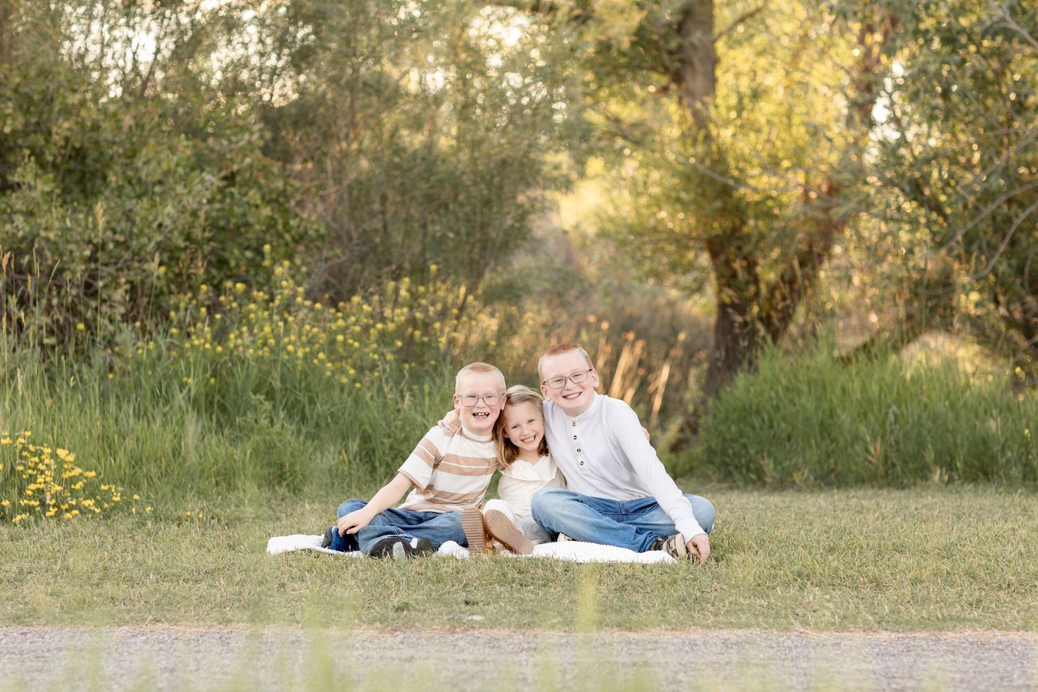 Three siblings hugging and smiling together on a blanket in a lush green outdoor setting.