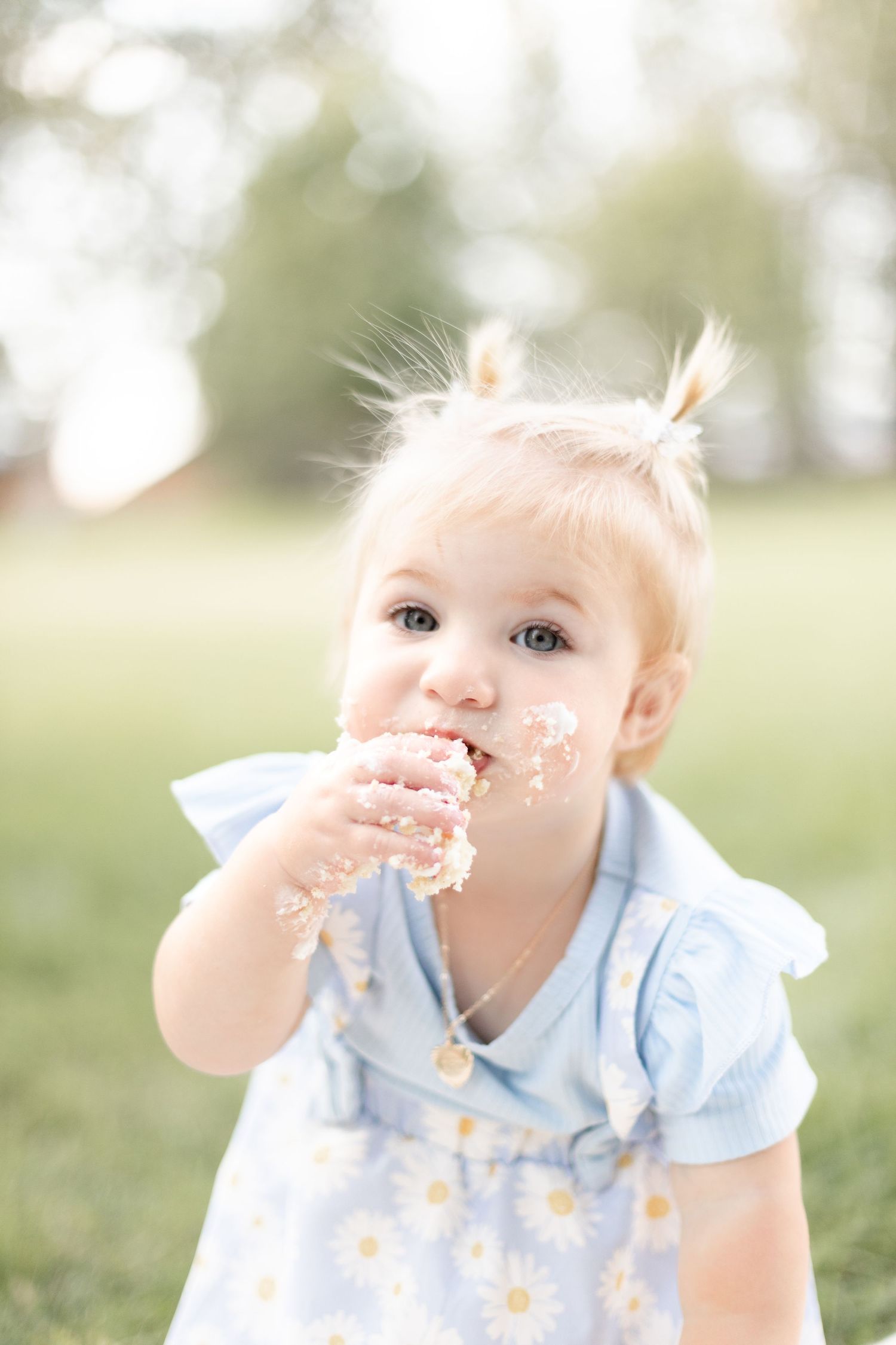 Cute baby girl with pigtails in blue floral dress enjoying cake smash outdoors in park.
