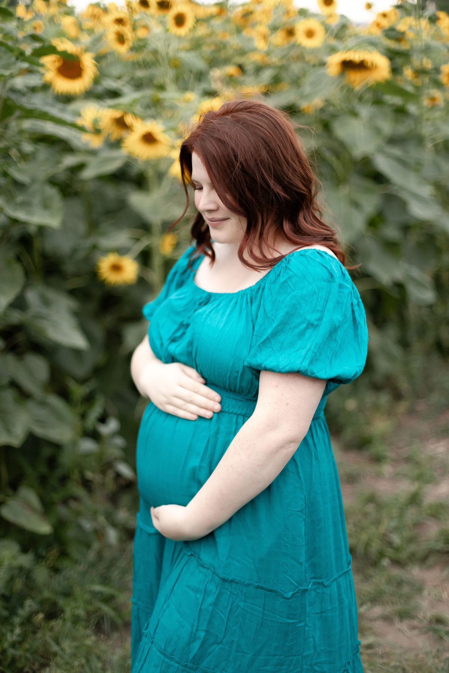 Pregnant woman in teal dress cradles baby bump while standing among tall sunflowers in soft light.