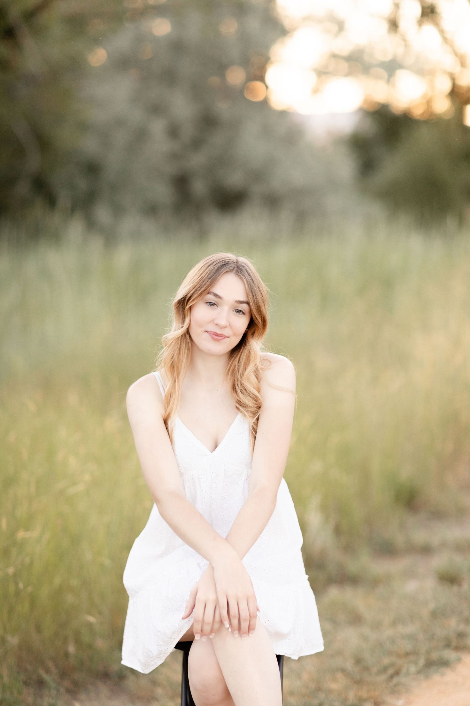Young woman in white sundress sitting outdoors in golden hour light with lush green field background.