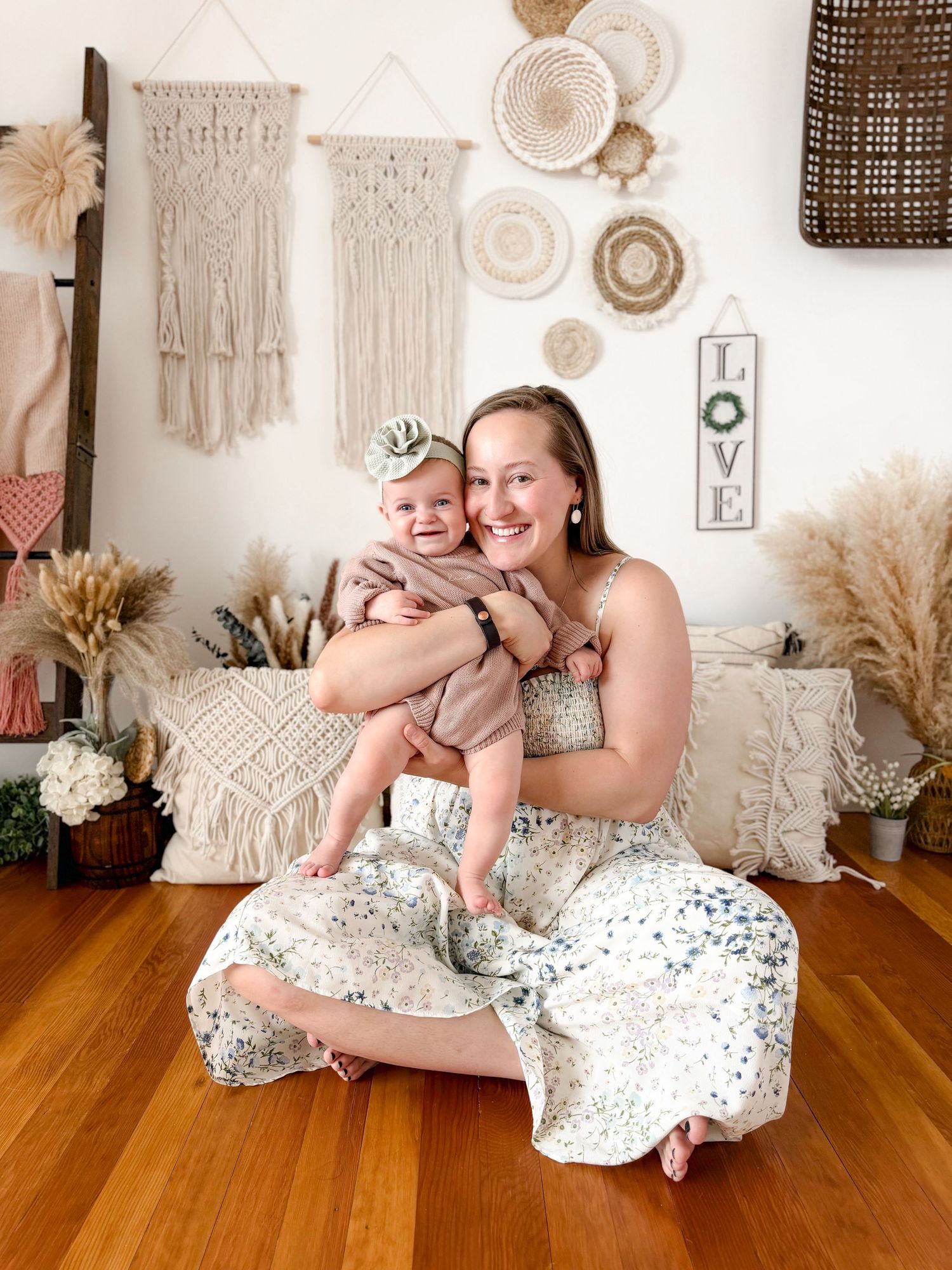 Smiling mother in floral dress holding baby girl in boho studio with macrame wall decor.