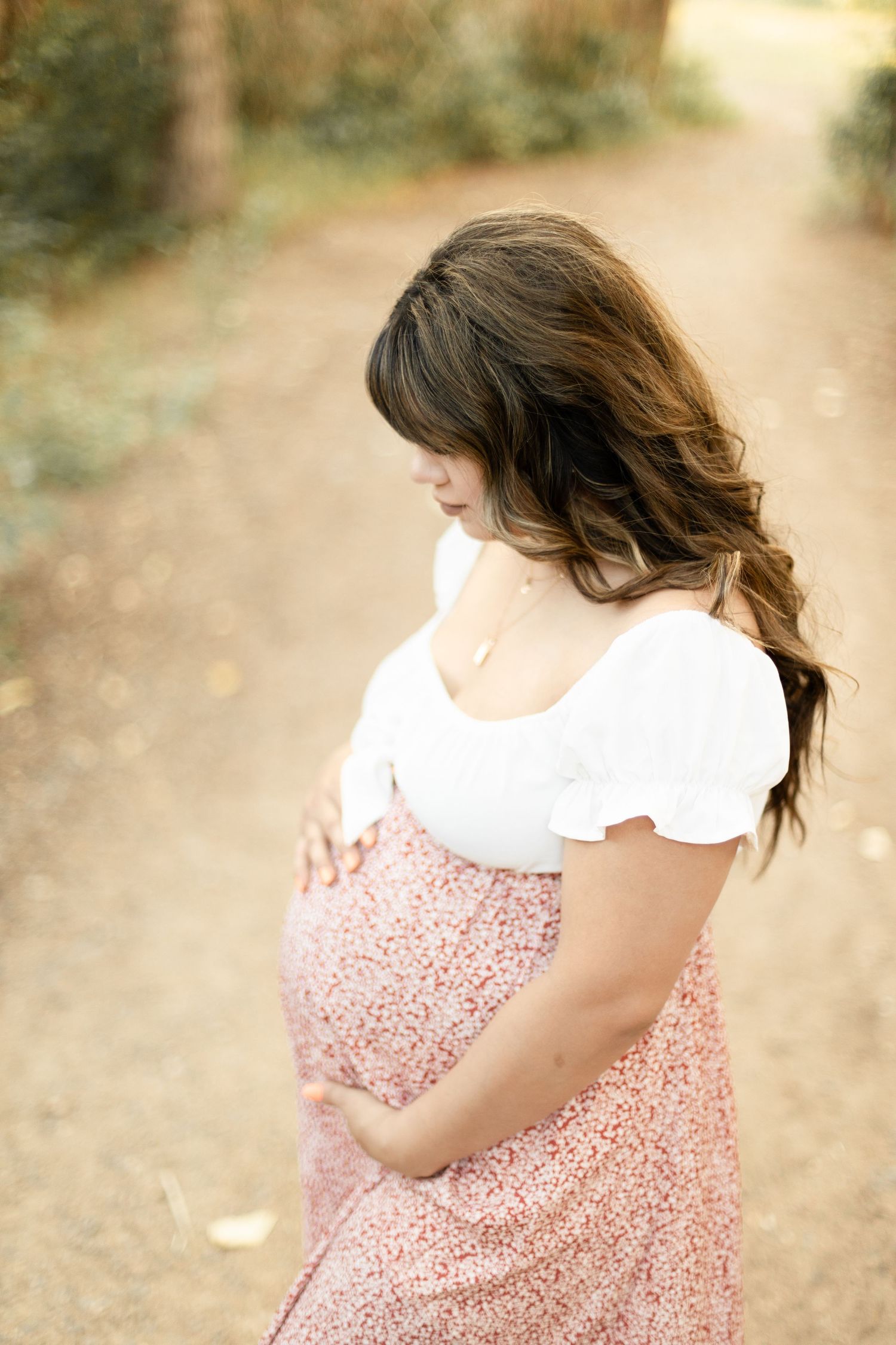 Pregnant brunette woman in white top and floral skirt cradles baby bump on a sunlit wooded path.