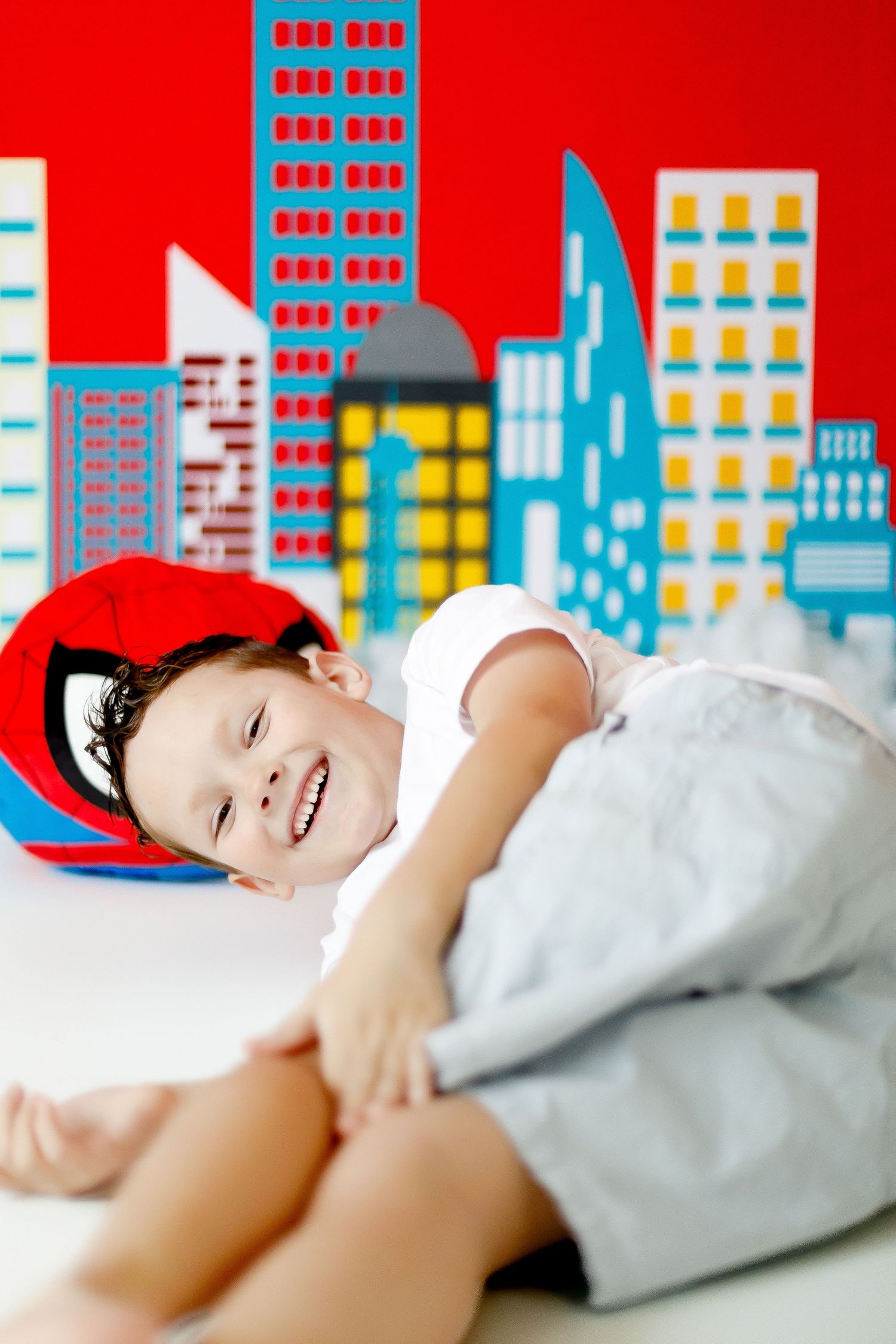 A smiling child wearing a Spider-Man hat lies down in front of a colorful superhero city backdrop.