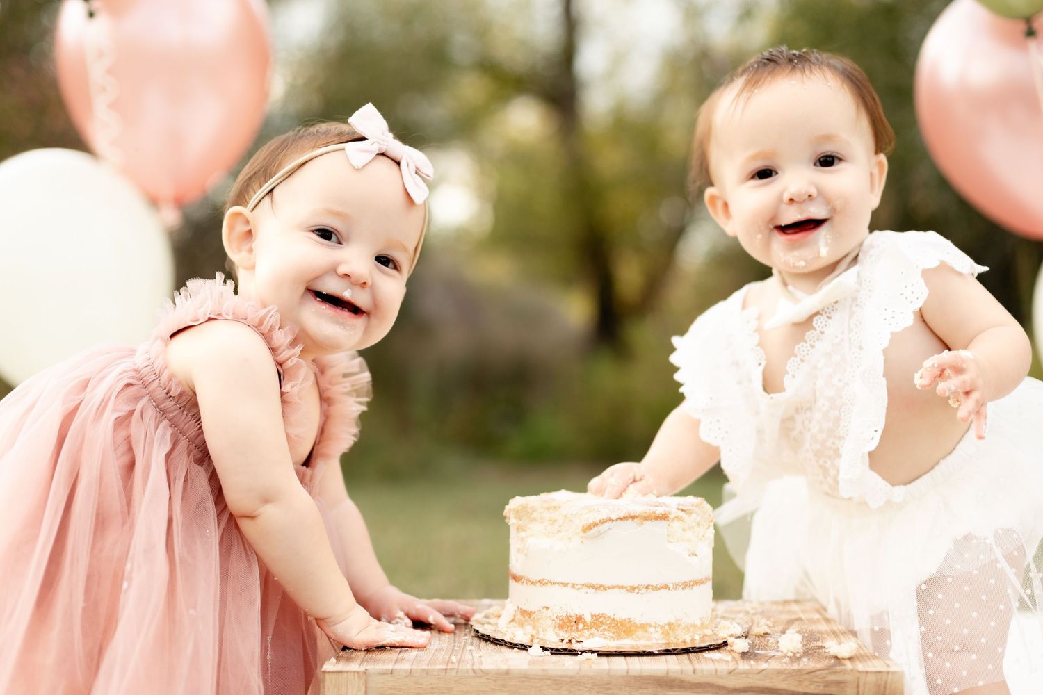 Two smiling baby girls in pink and white dresses celebrating first birthday with cake smash outdoors with balloons.
