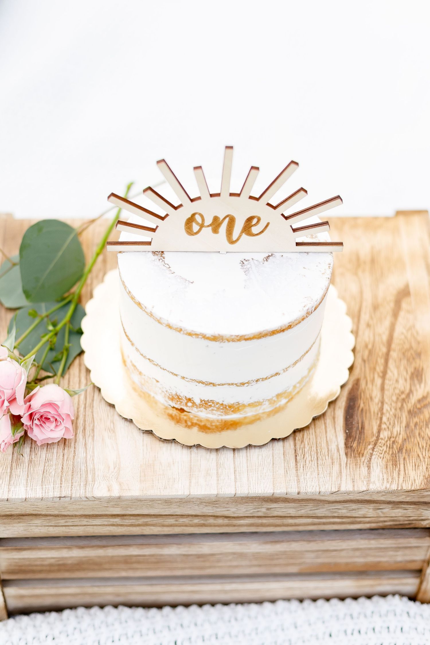 Elegant first birthday cake with gold sun topper reading one on wooden crate with pink roses.