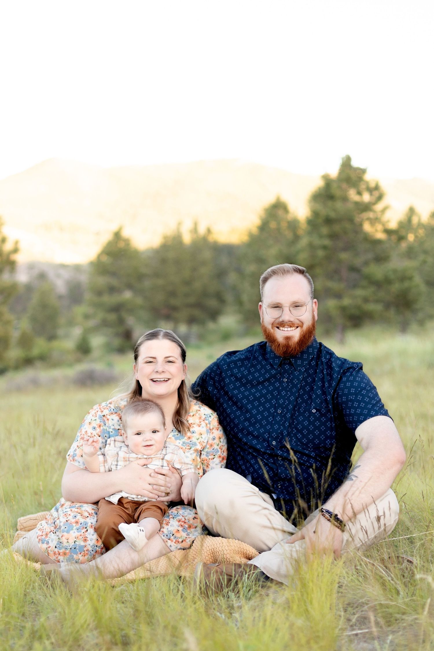 Young family with baby sitting in tall grass with mountain backdrop at golden hour.