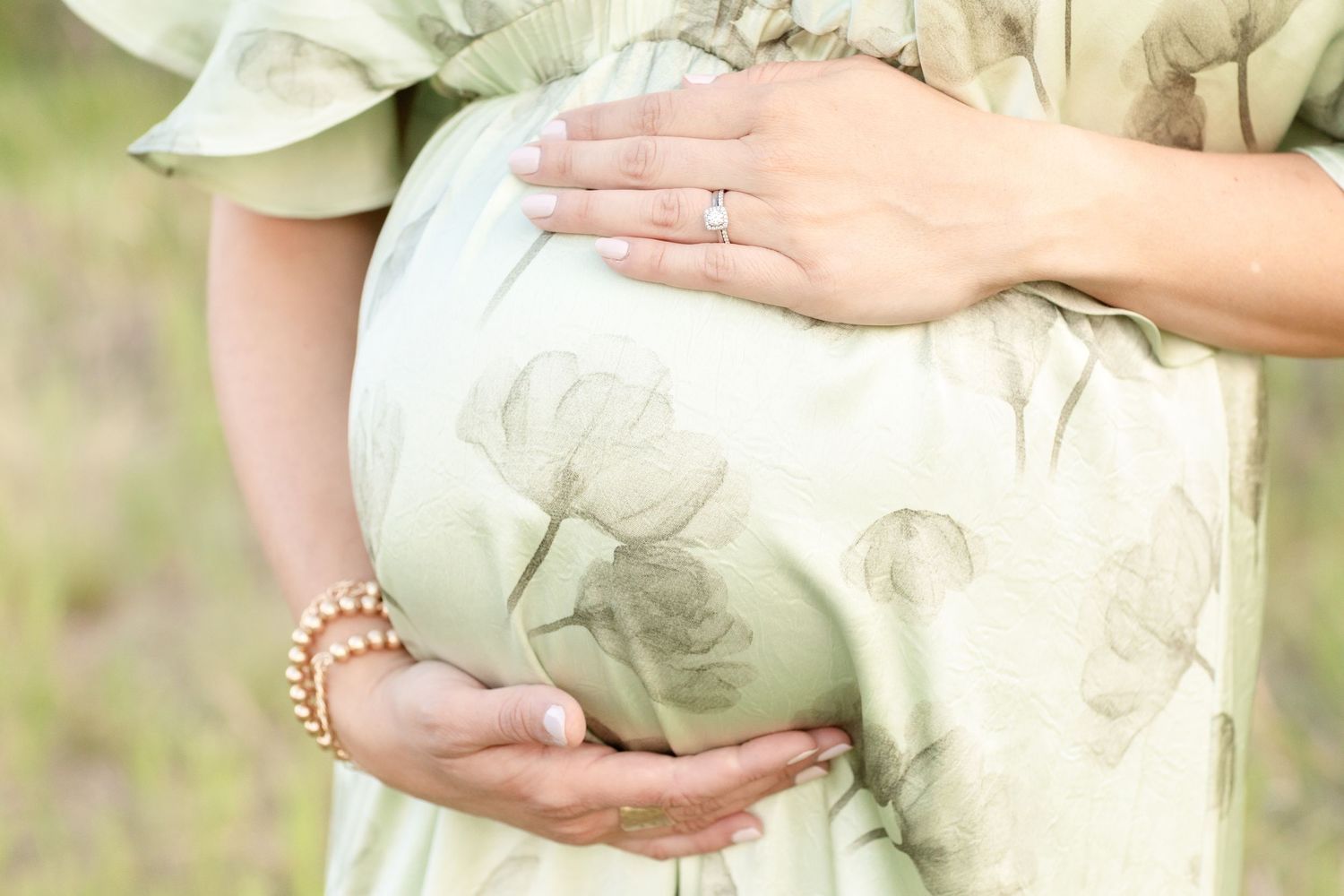 Close-up of pregnant woman in sage green floral dress cradling baby bump with ring and pearl bracelet.