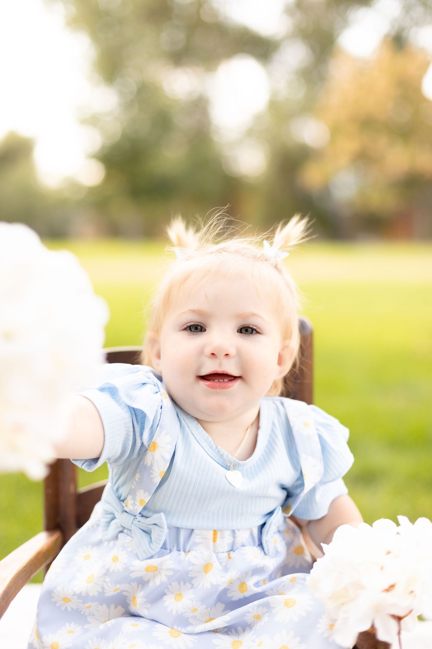 Cheerful baby girl in a blue daisy dress reaching toward white flowers while sitting in a wooden chair outdoors.