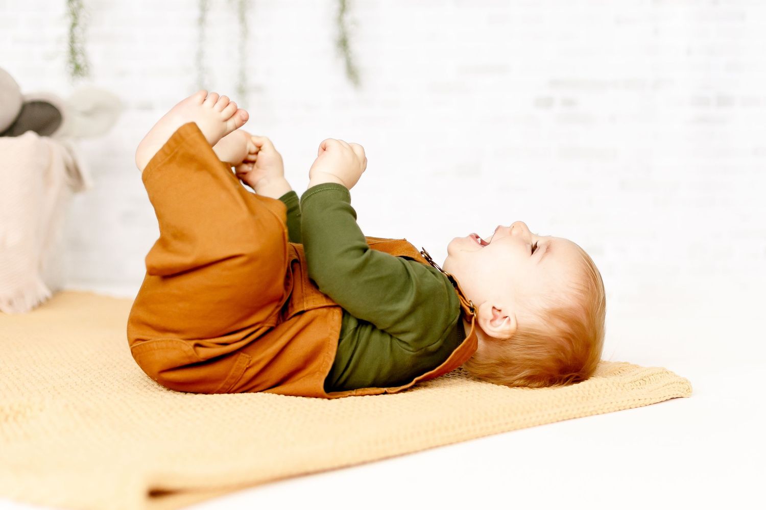 A happy baby lying on their back on a yellow blanket, grabbing their feet while wearing an orange and green outfit.