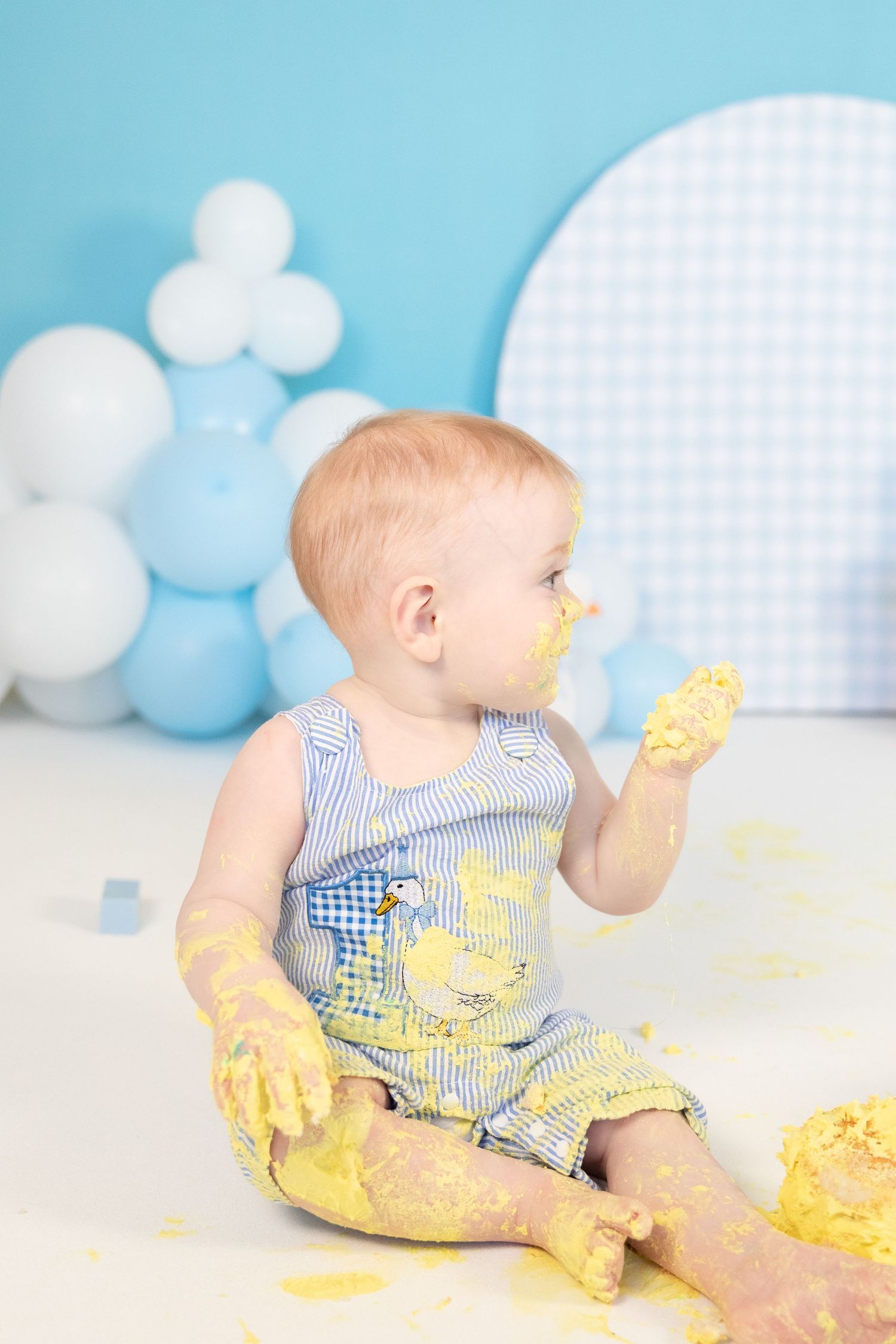 Baby in blue romper eating yellow cake with hands during first birthday cake smash photoshoot.