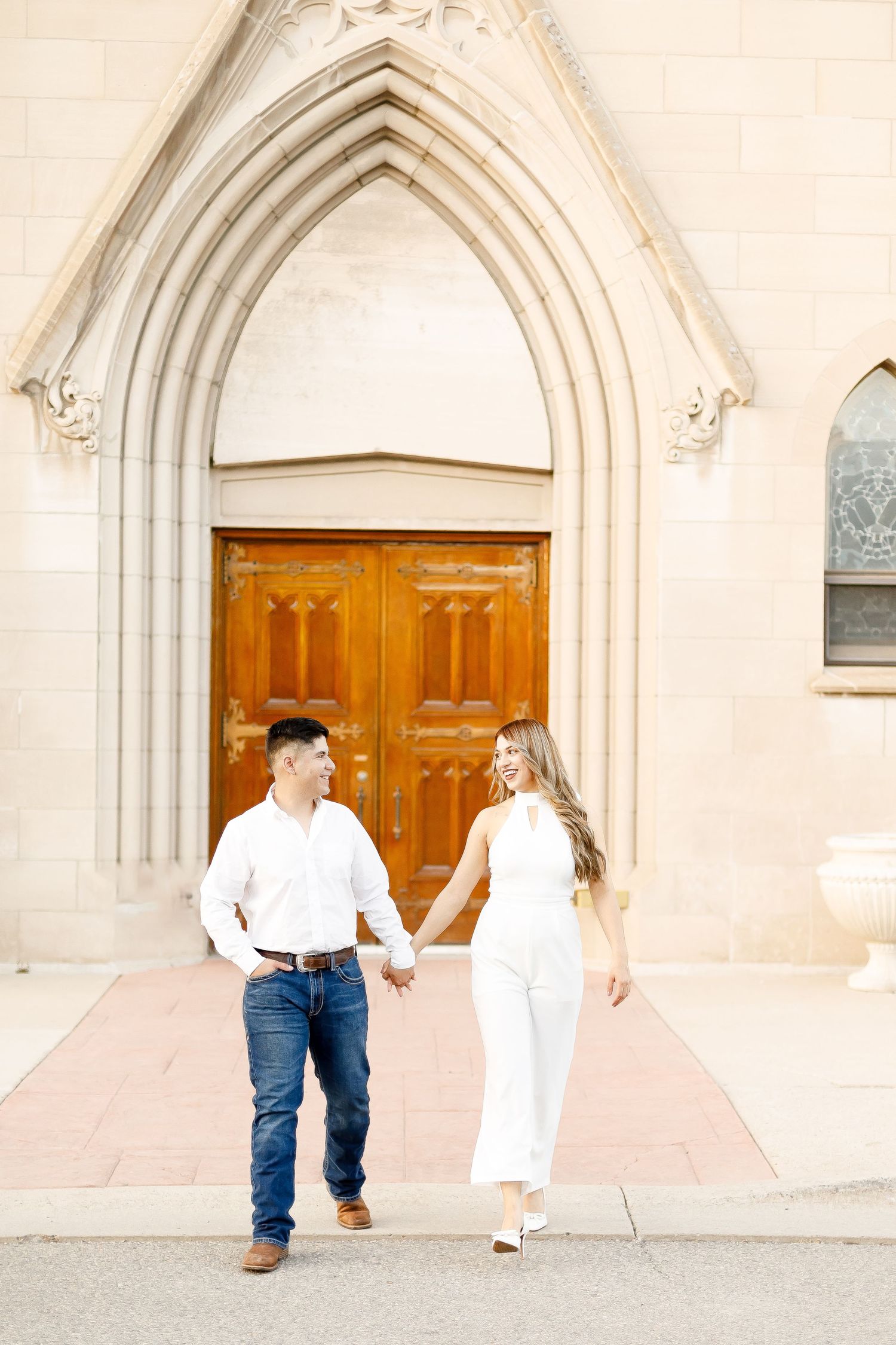 Couple in white outfits holding hands walking in front of gothic church entrance.