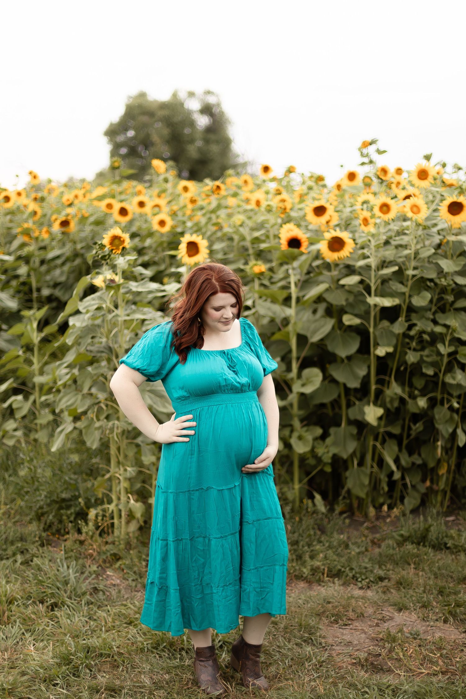 Pregnant woman in teal dress and brown boots poses thoughtfully in a lush sunflower field.