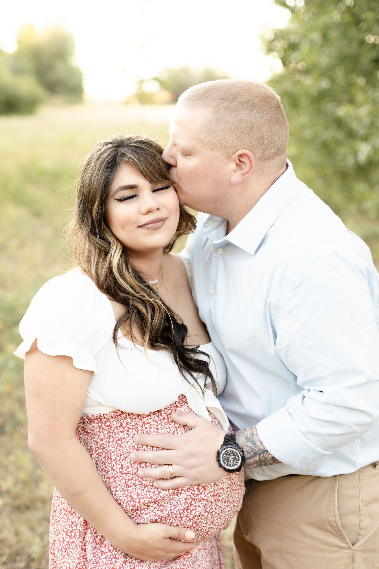 Man tenderly kisses pregnant woman on cheek while cradling her baby bump during a sunny outdoor maternity session.
