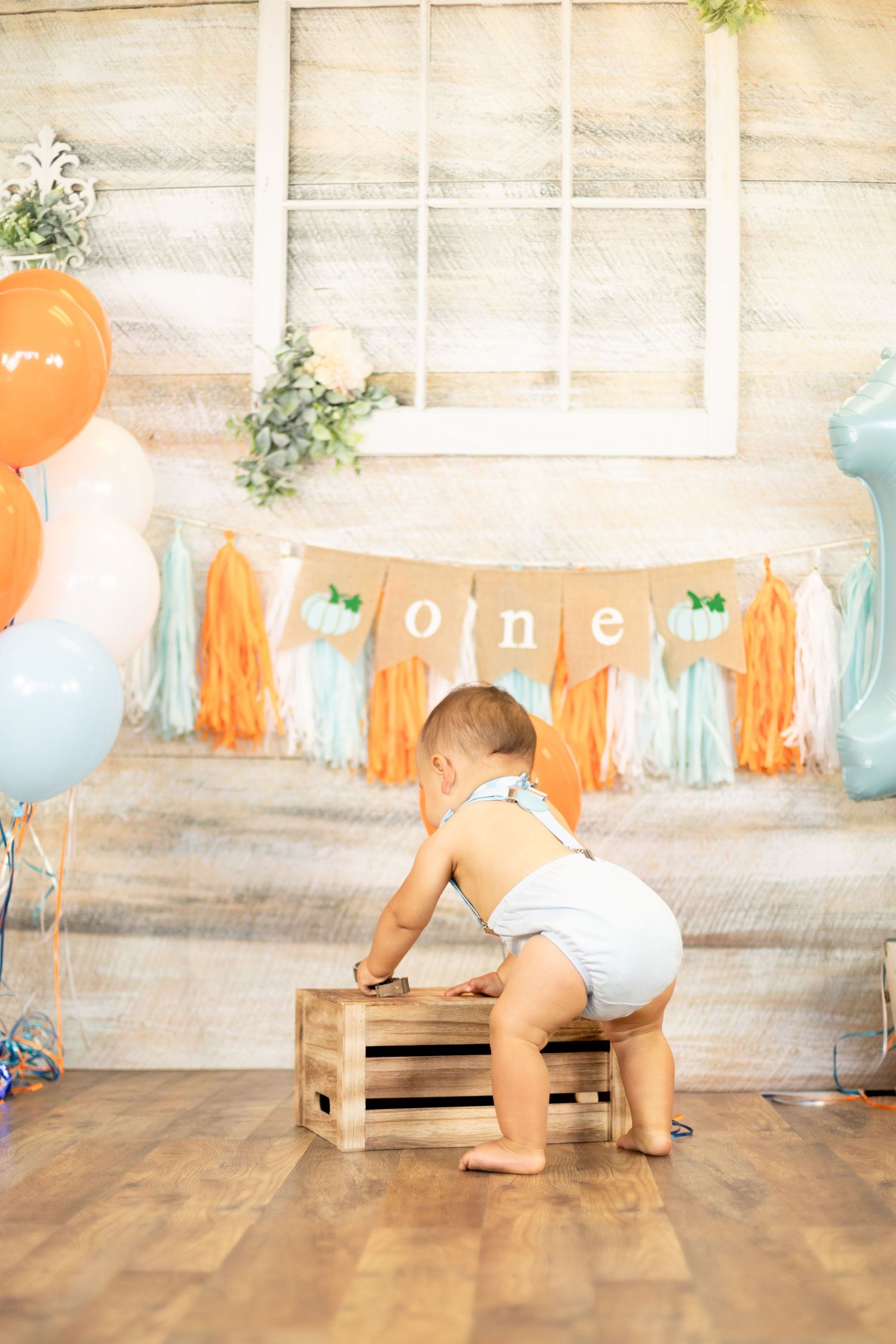 Baby boy in white diaper with suspenders climbing on wooden crate at first birthday party with one banner.
