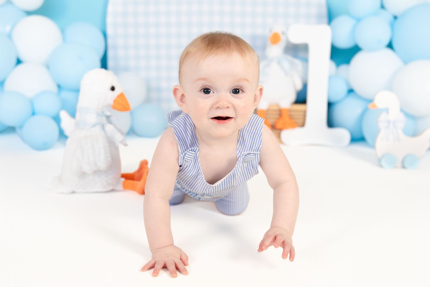 Bright-eyed baby boy in striped romper crawls on white floor with duck and balloon birthday decorations.