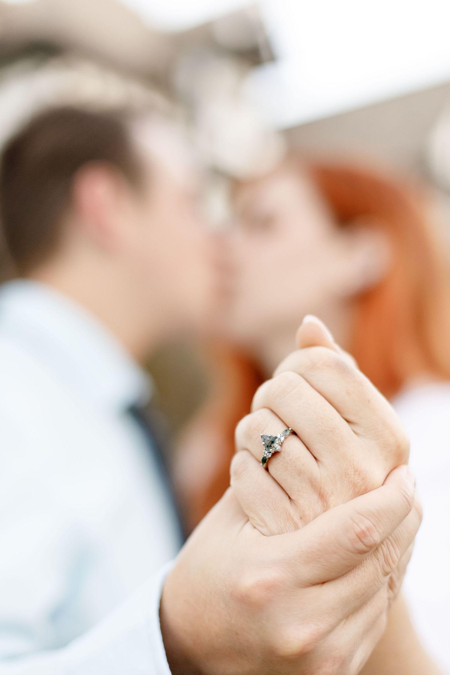 Close-up of an engagement ring on a woman's hand while a couple kisses blurred in the background.