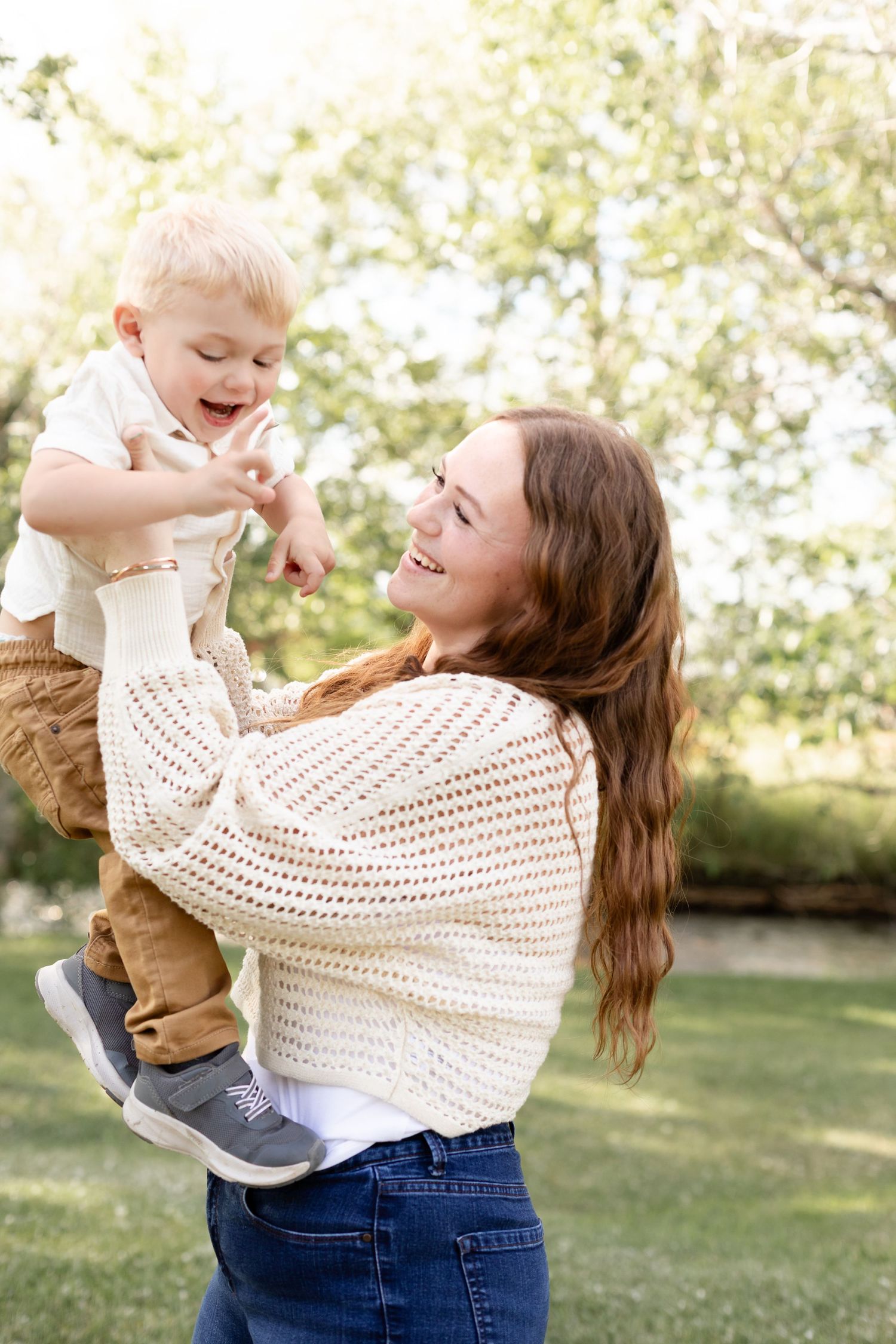 Smiling mother lifting her laughing toddler son in a sunny outdoor park setting.