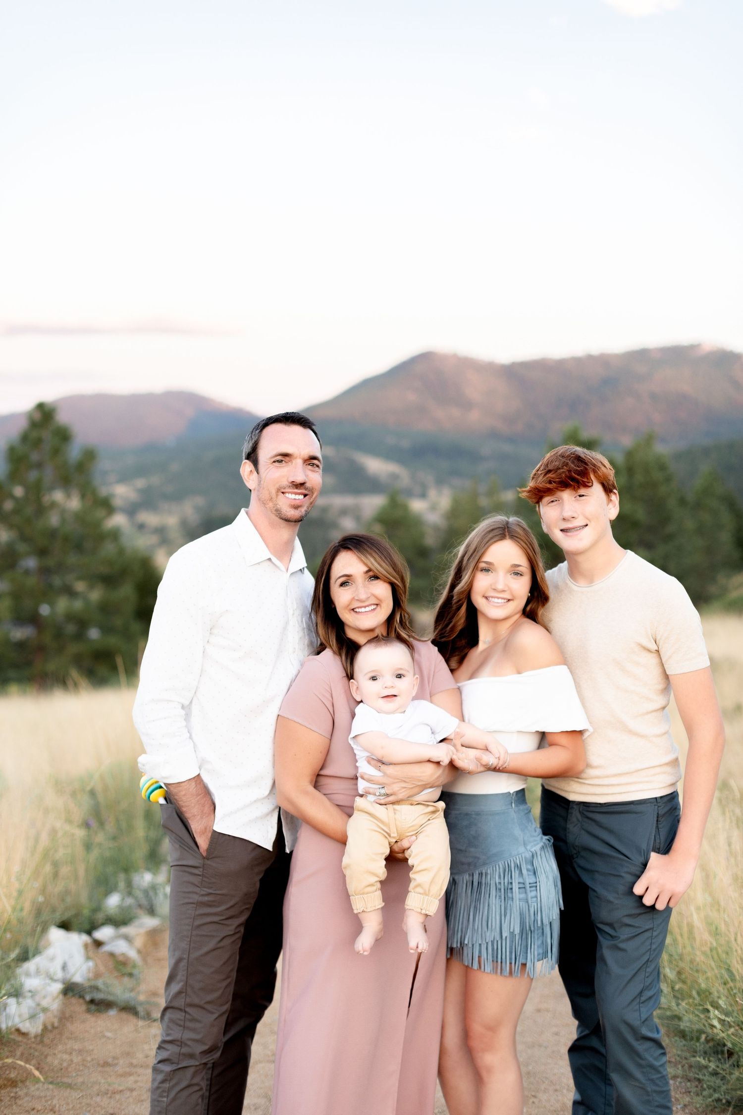 A family of five poses together on a rocky path with mountain and pine tree scenery at sunset.