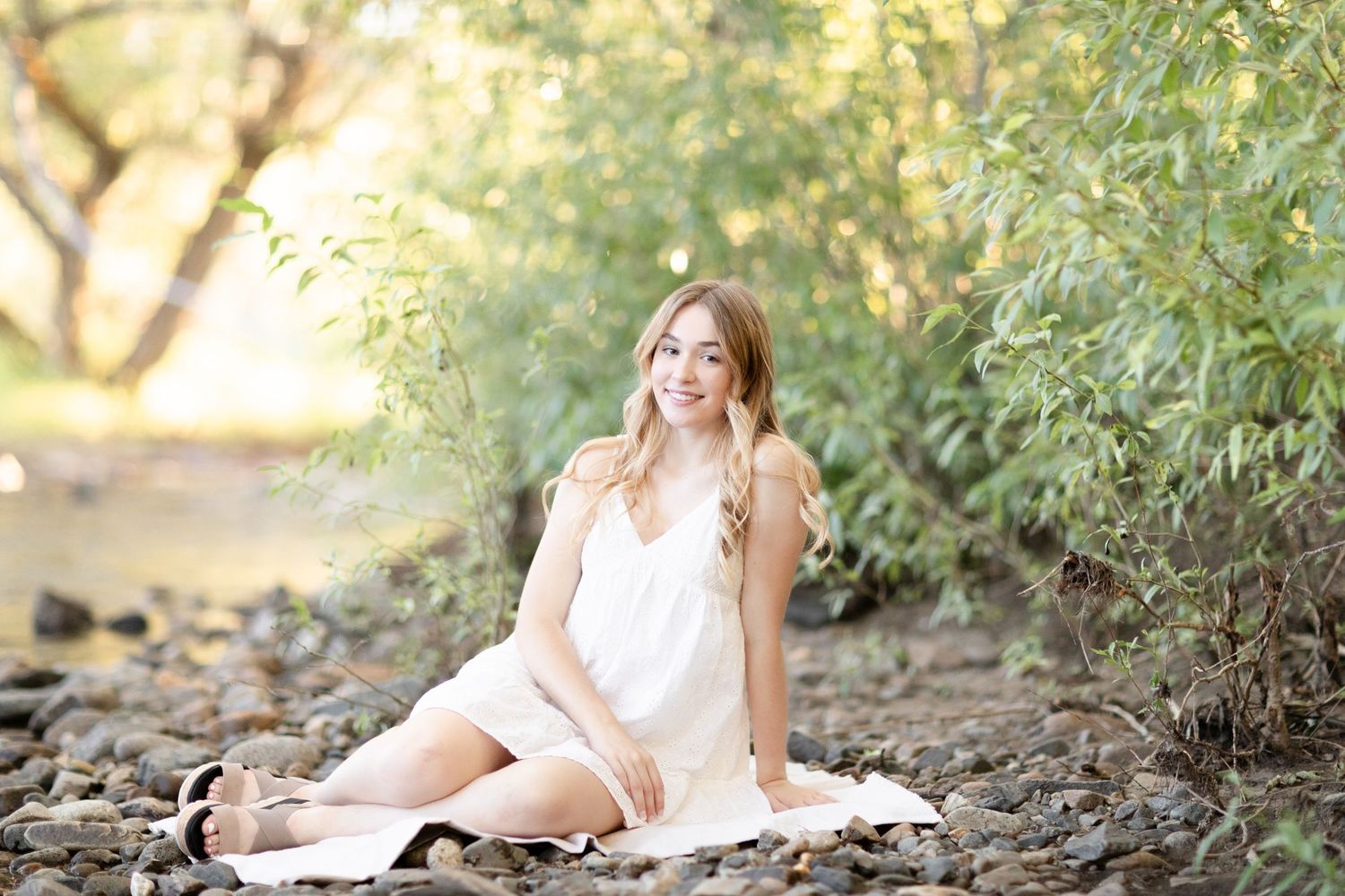 Young woman in white dress sitting on rocky ground near lush green trees in a serene outdoor setting.