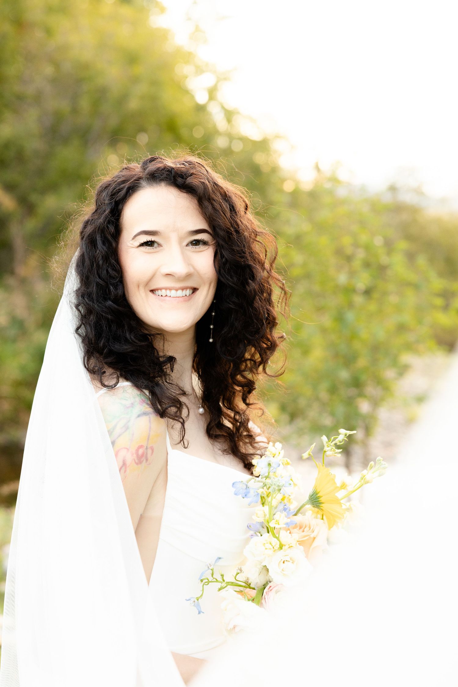 Smiling bride with curly hair holding yellow and blue wildflower bouquet outdoors.