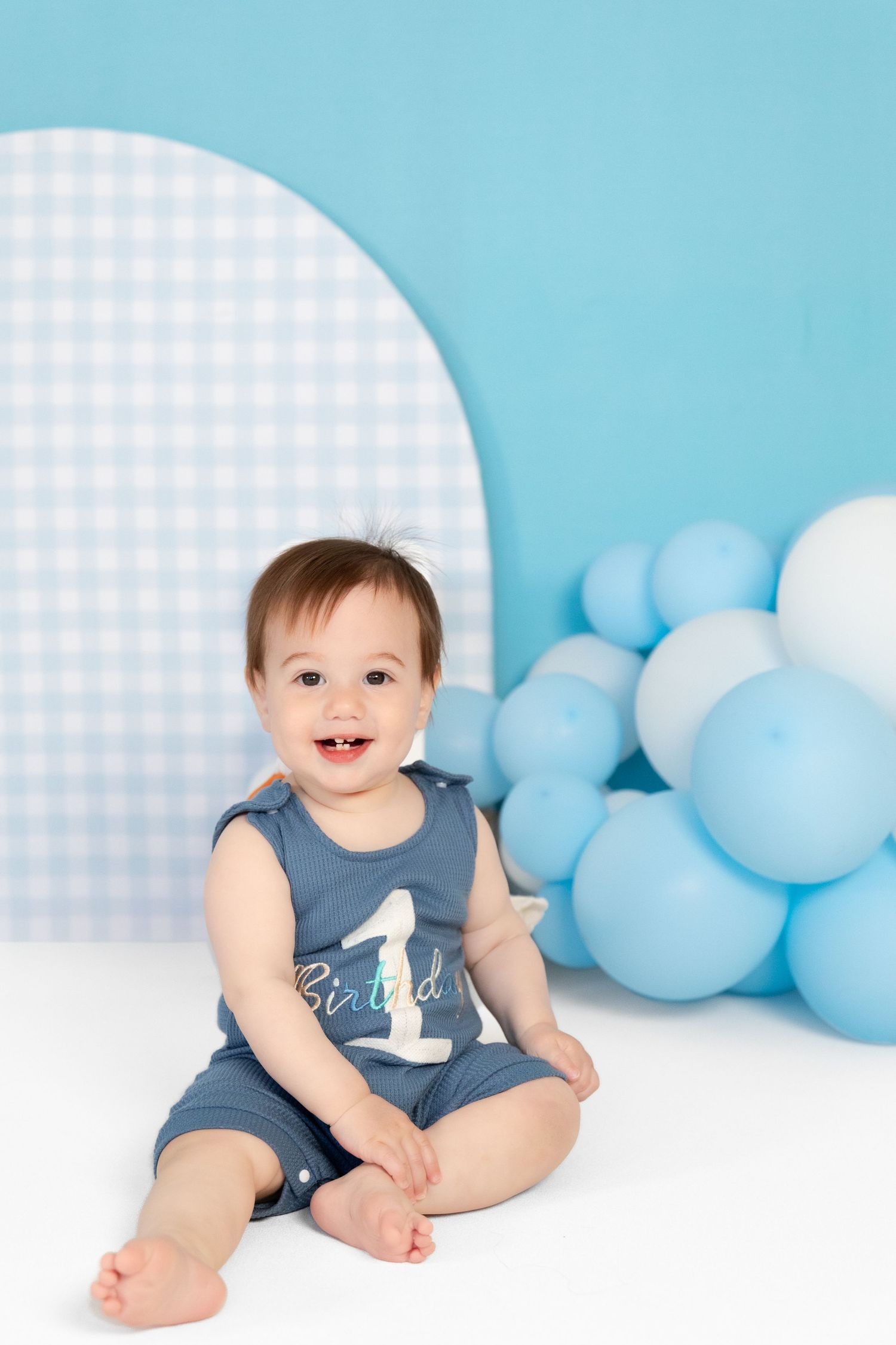 Smiling baby boy in blue romper sitting with balloons at duck-themed first birthday photo session.