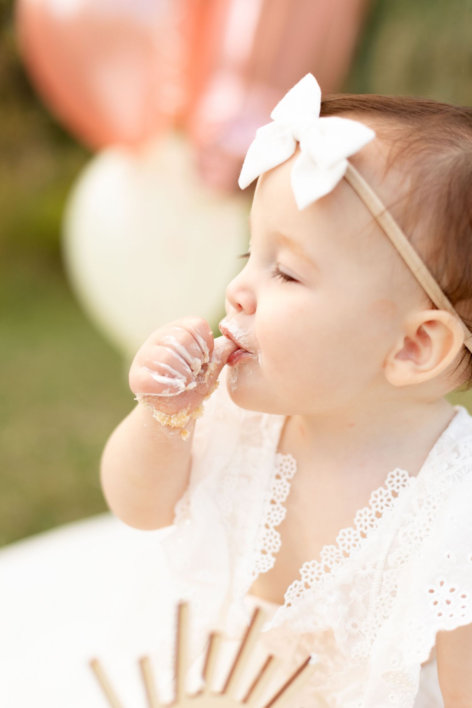 Baby girl in white lace dress and bow headband eating cake at outdoor first birthday party with pink balloons.