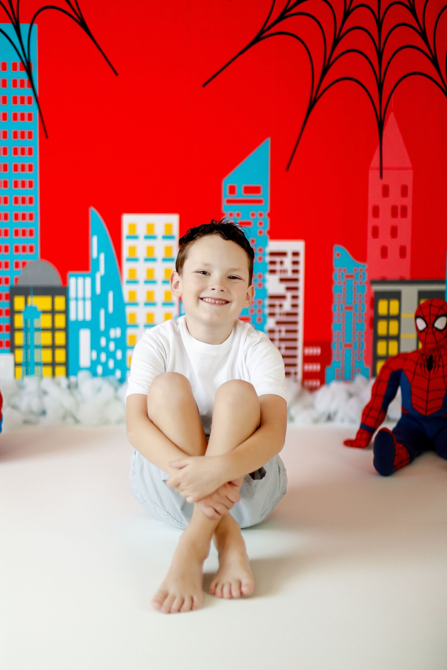 A smiling boy sits cross-legged in front of a vibrant Spider-Man themed birthday party backdrop.